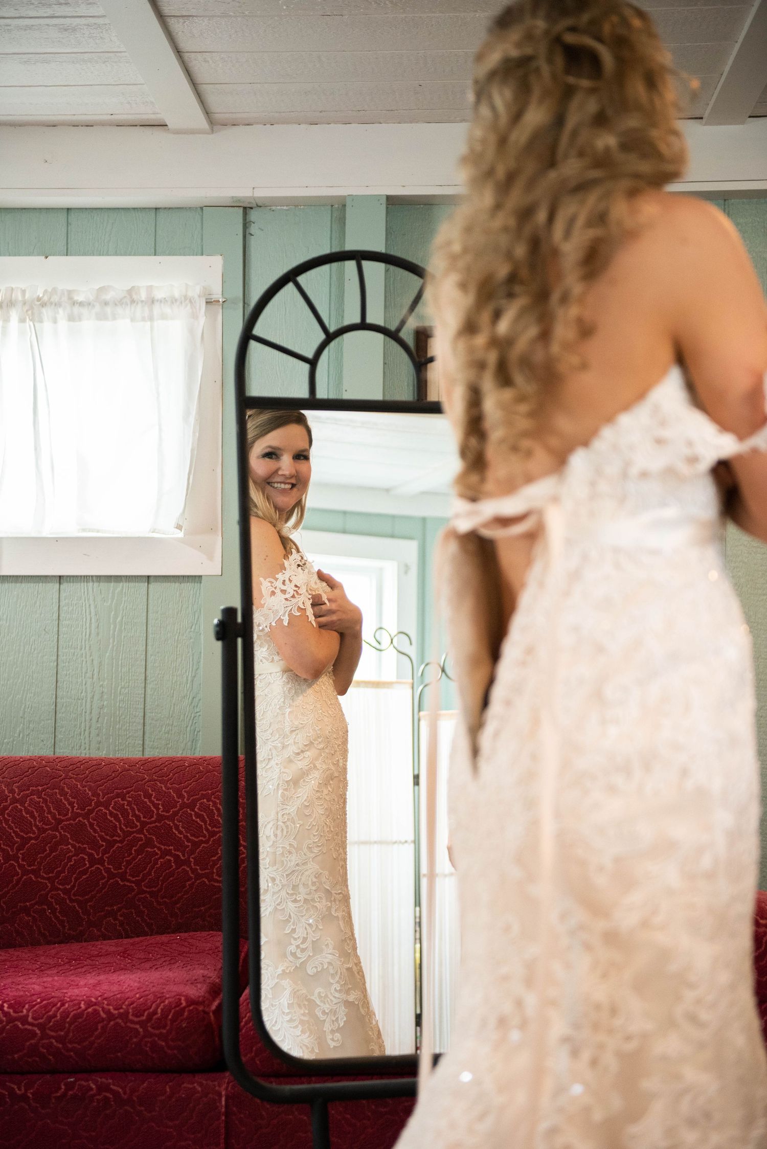 A bride admires her reflection in a full-length mirror while wearing a fitted lace wedding gown.