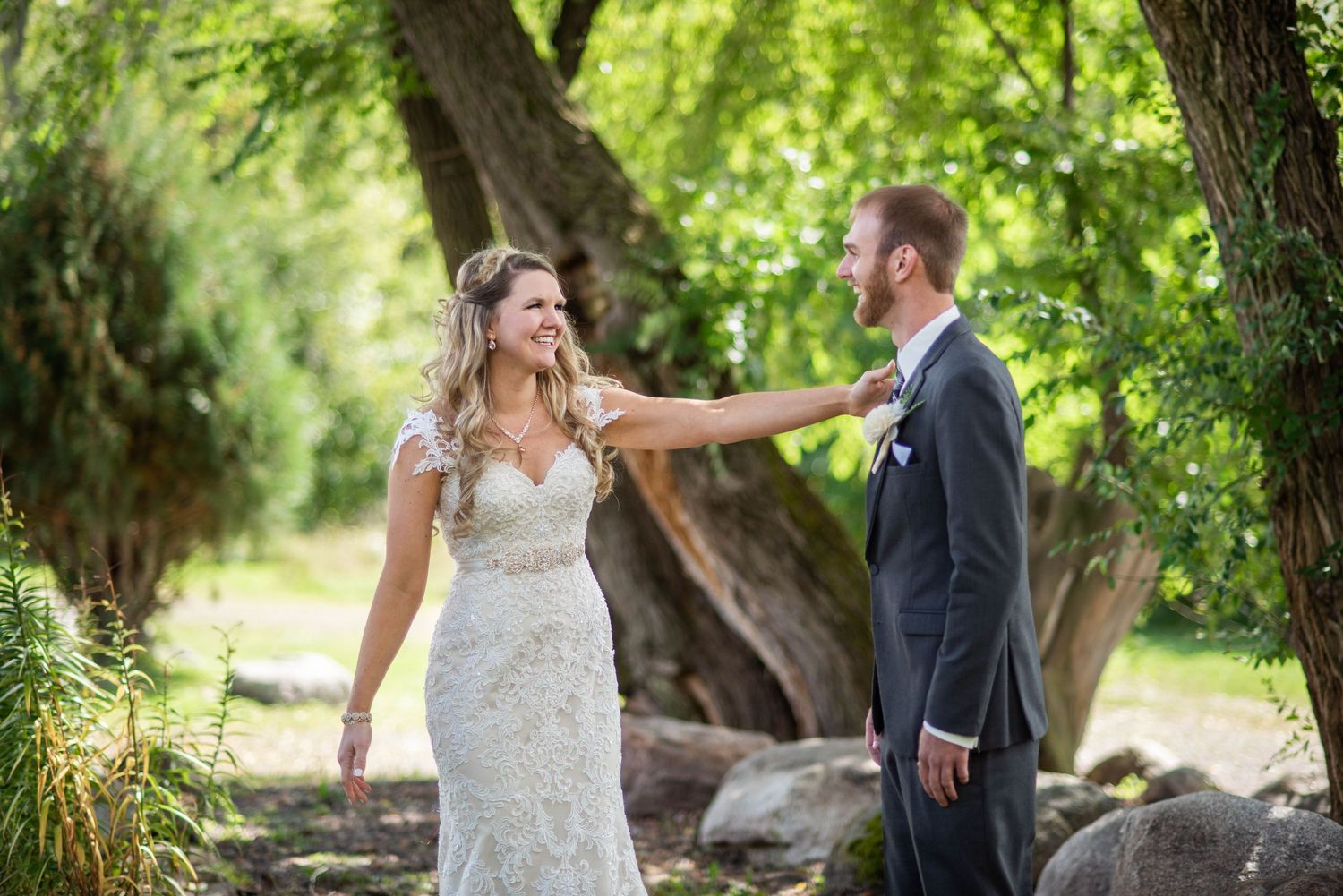 A couple shares a joyful moment on their wedding day in a lush garden setting with green trees in the background.