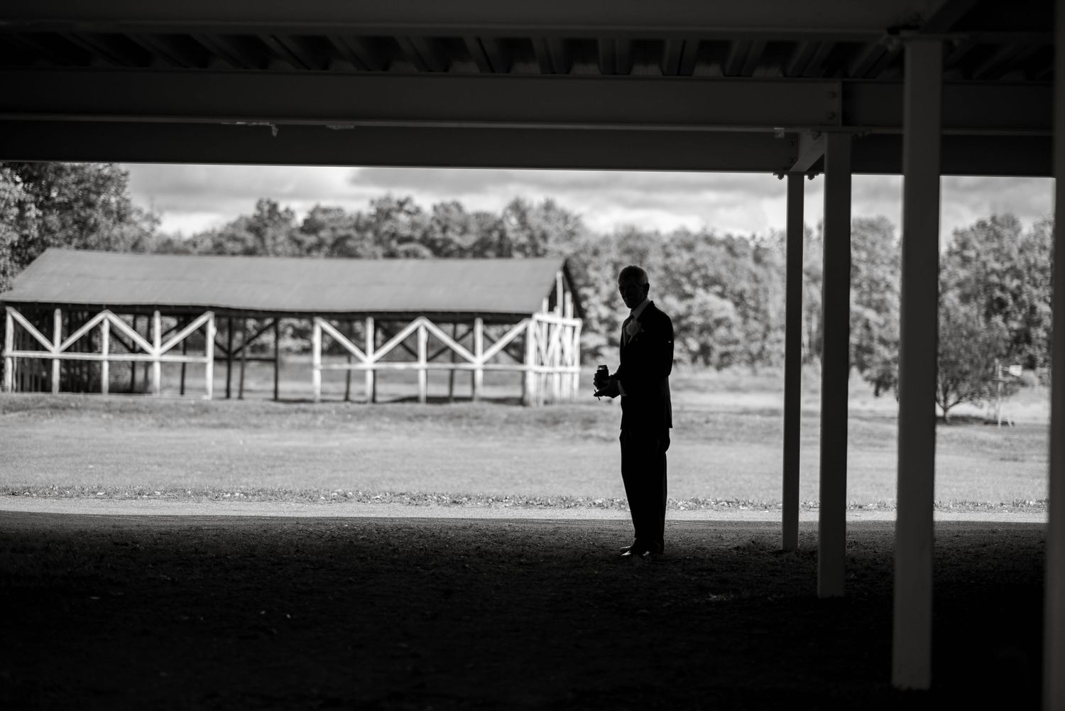 A silhouetted figure stands at the edge of a covered area overlooking a fence and fields in black and white photography.
