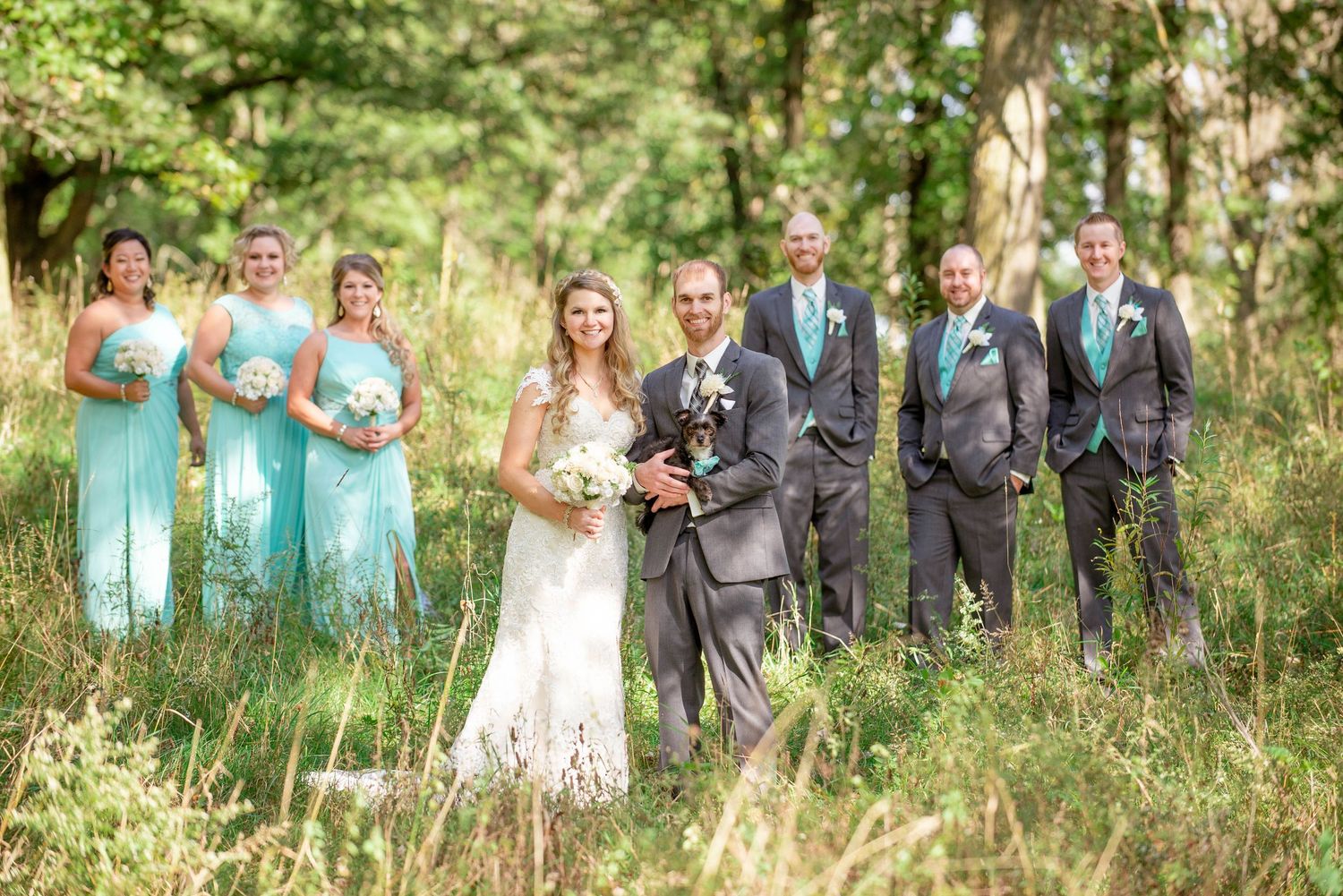A wedding party with bridesmaids in mint dresses and groomsmen pose together in a wooded setting. Rum River Barn Milaca Minnesota Weddings