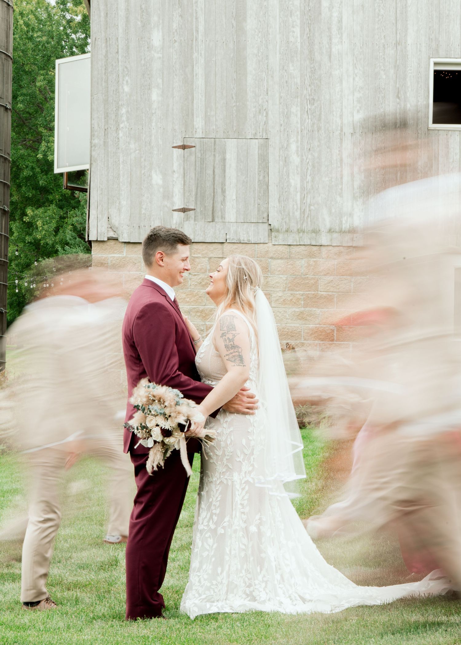 A couple in wedding attire embraces outdoors by a rustic barn wall while motion-blurred figures pass around them.