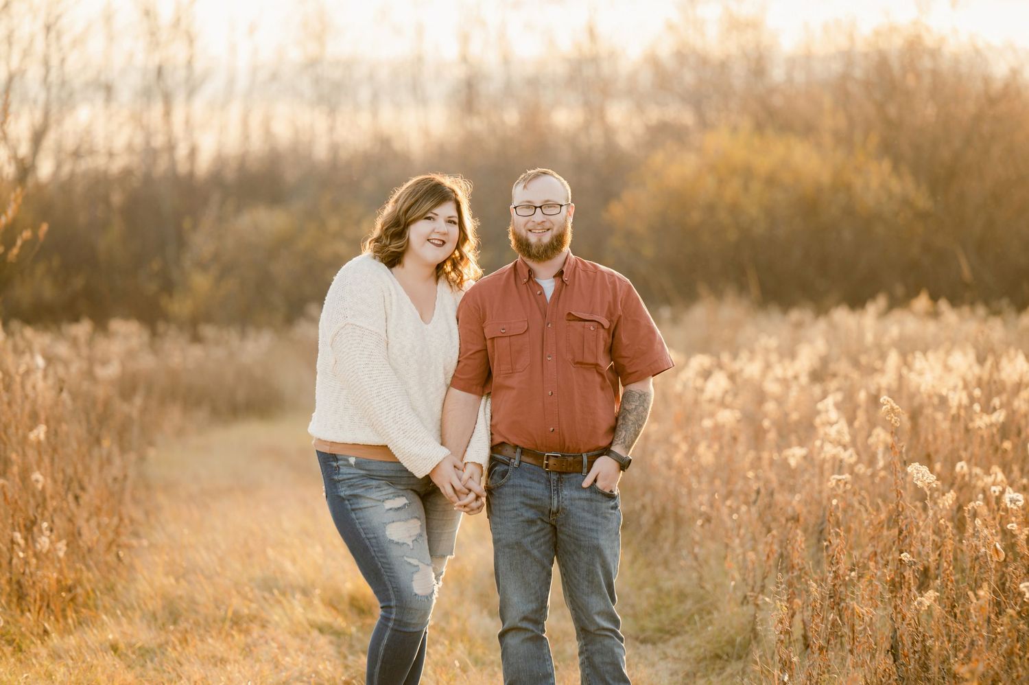 A couple walks hand in hand on a dirt path through golden grass during sunset.