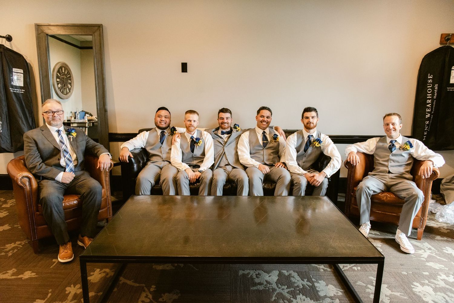 Groomsmen in matching gray suits sitting on a wooden bench at a rustic wedding venue.