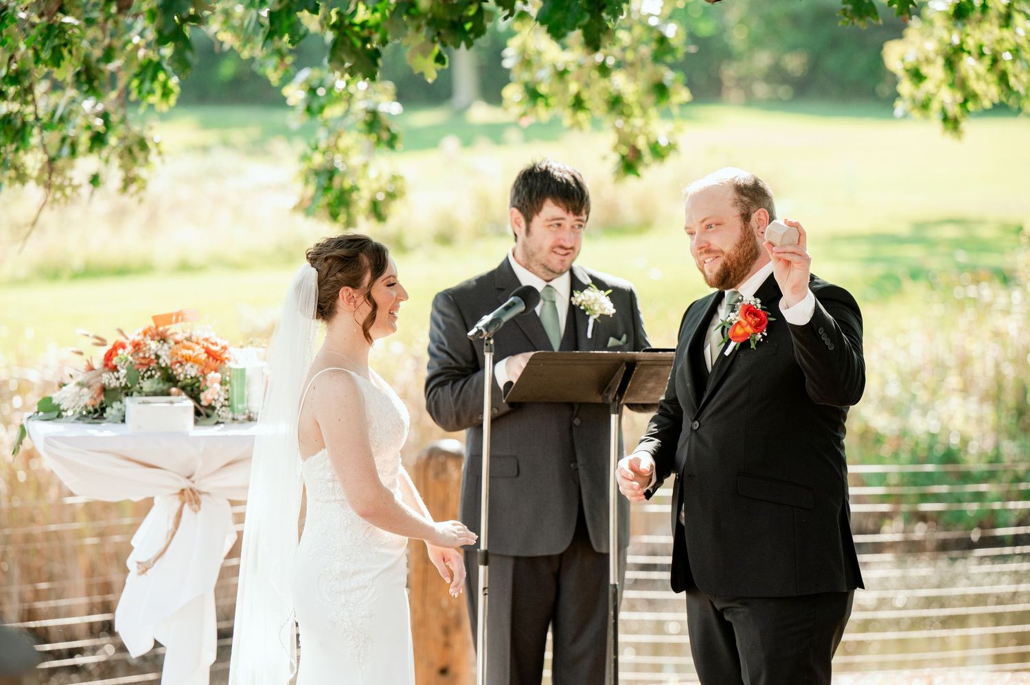 Outdoor wedding ceremony with attendees gathered under green leafy trees.
