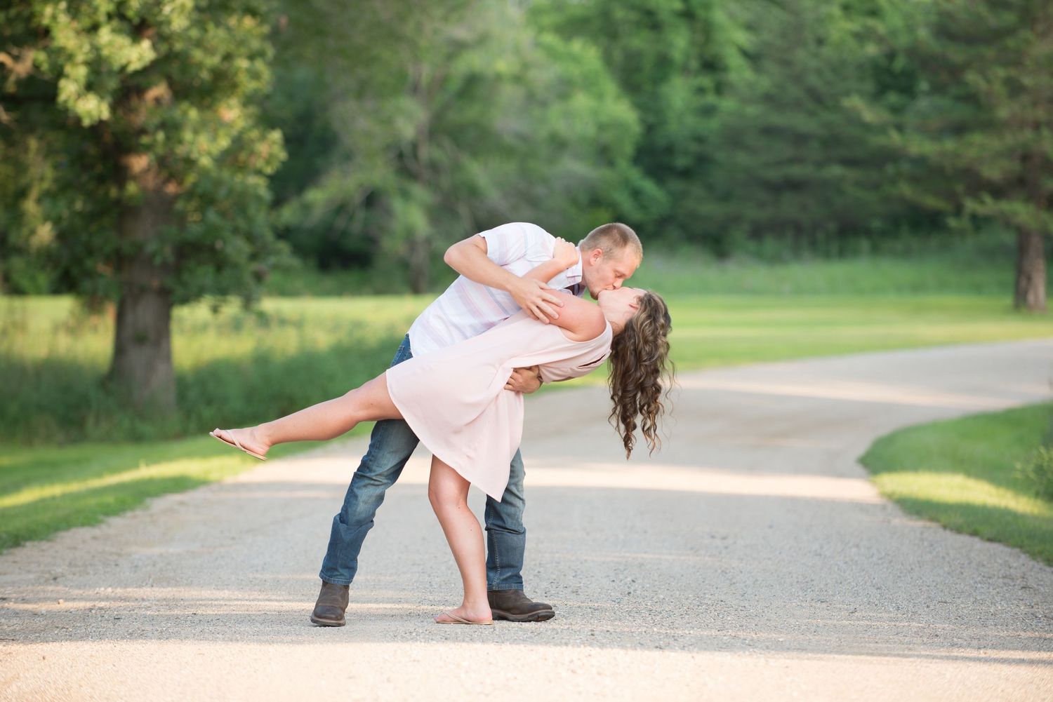 A romantic couple dancing together along a tree-lined path, creating a graceful dip pose in warm sunlight.