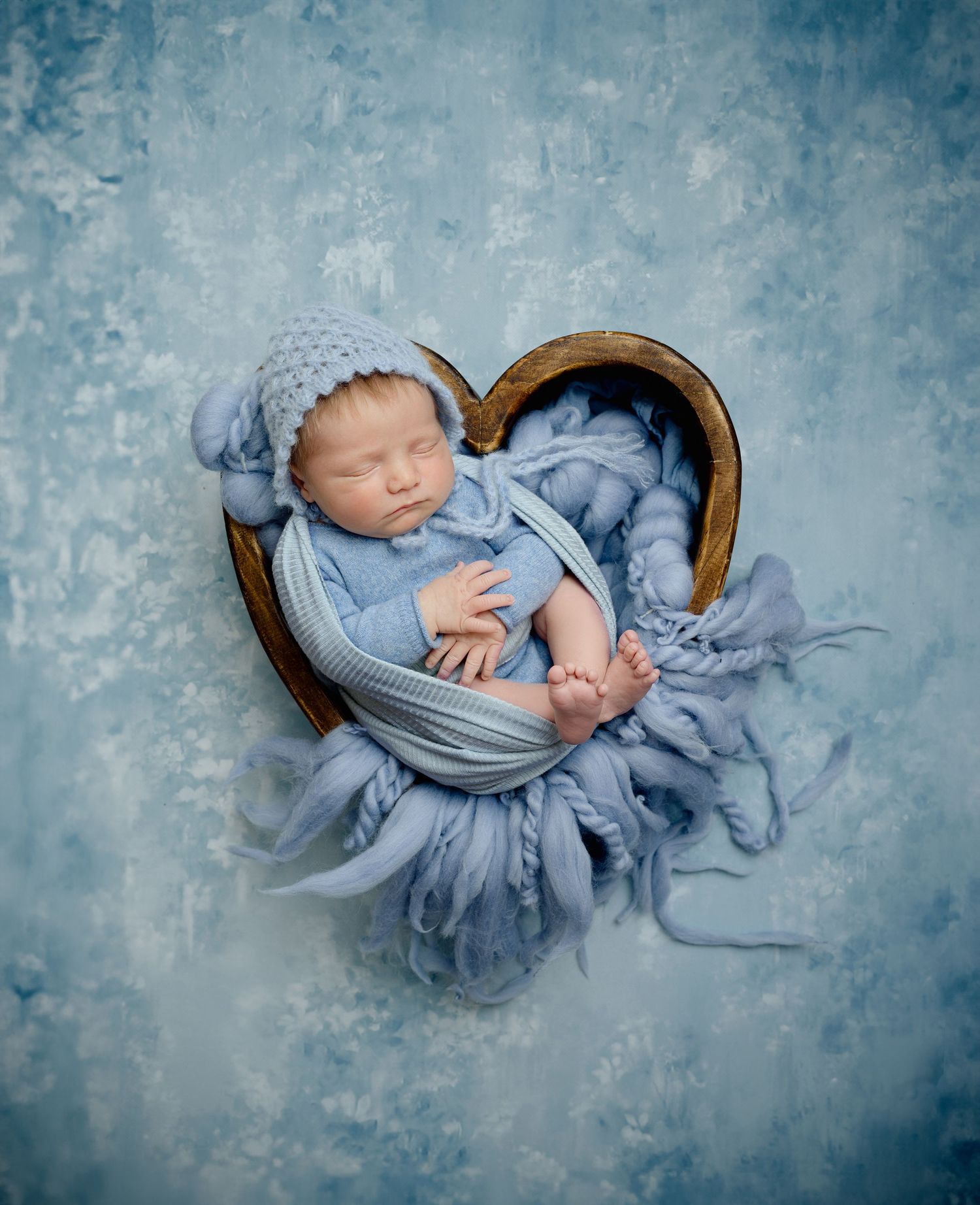 Newborn baby sleeping peacefully in a wooden bowl wrapped in soft blue fabric against a textured blue background.
