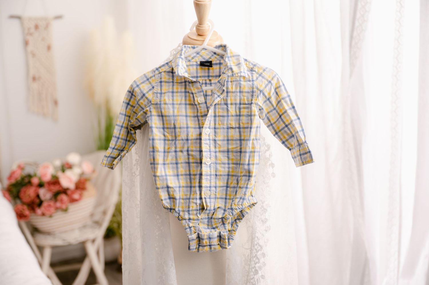 Baby plaid button-down onesie hangs from hanger near white curtains and floral decor.