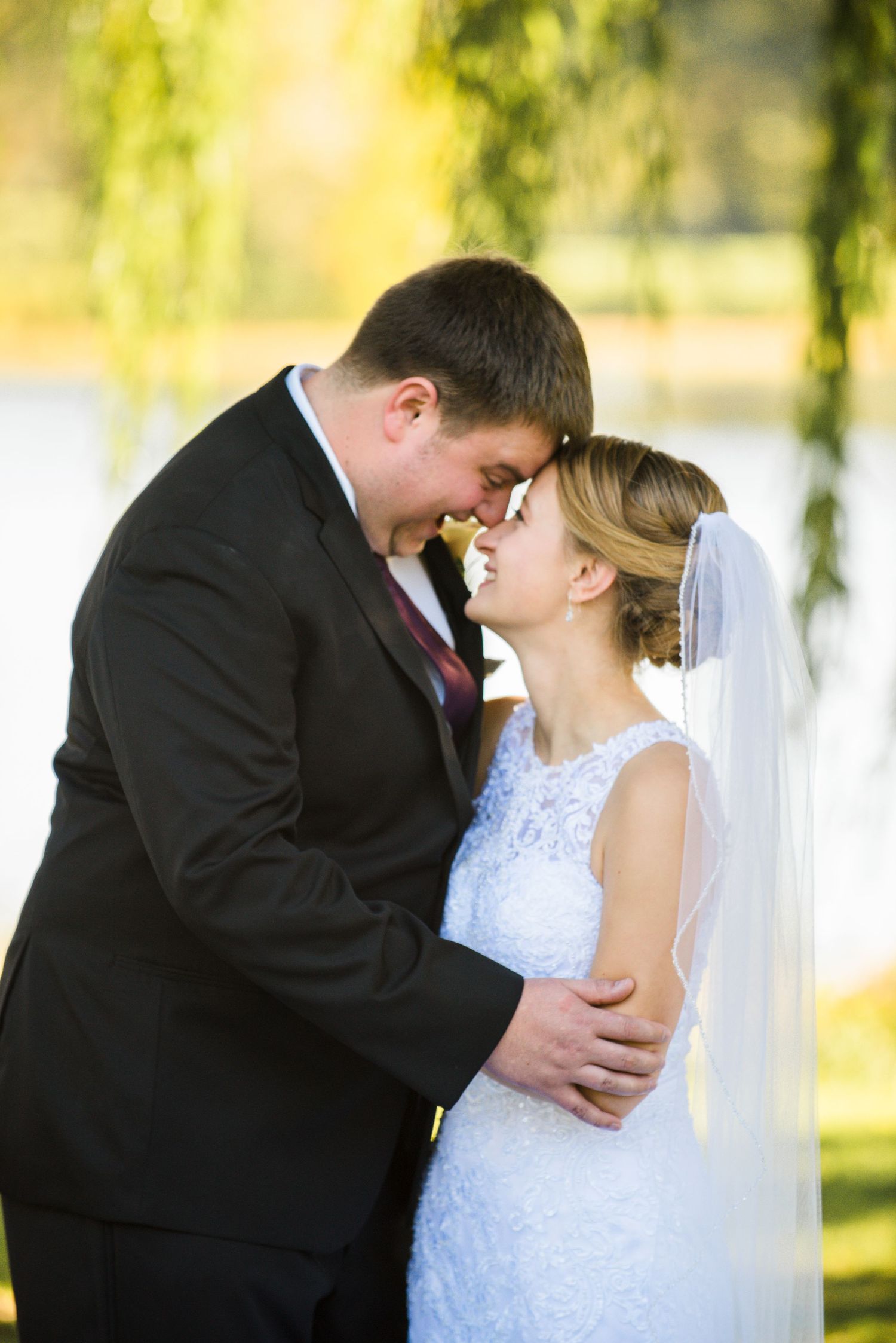 A tender wedding moment between newlyweds in formal attire beneath golden sunlit willow branches. Andover wedding at Magestic Oaks Golf Club
