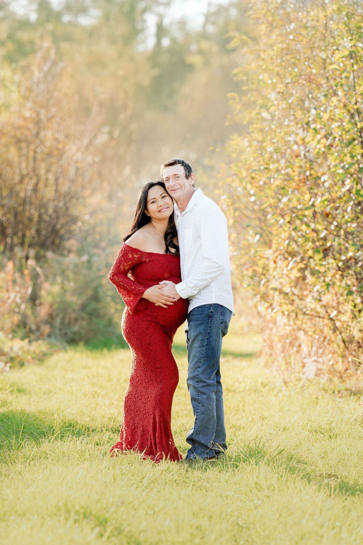Expecting couple in red dress and white shirt pose in autumn field.