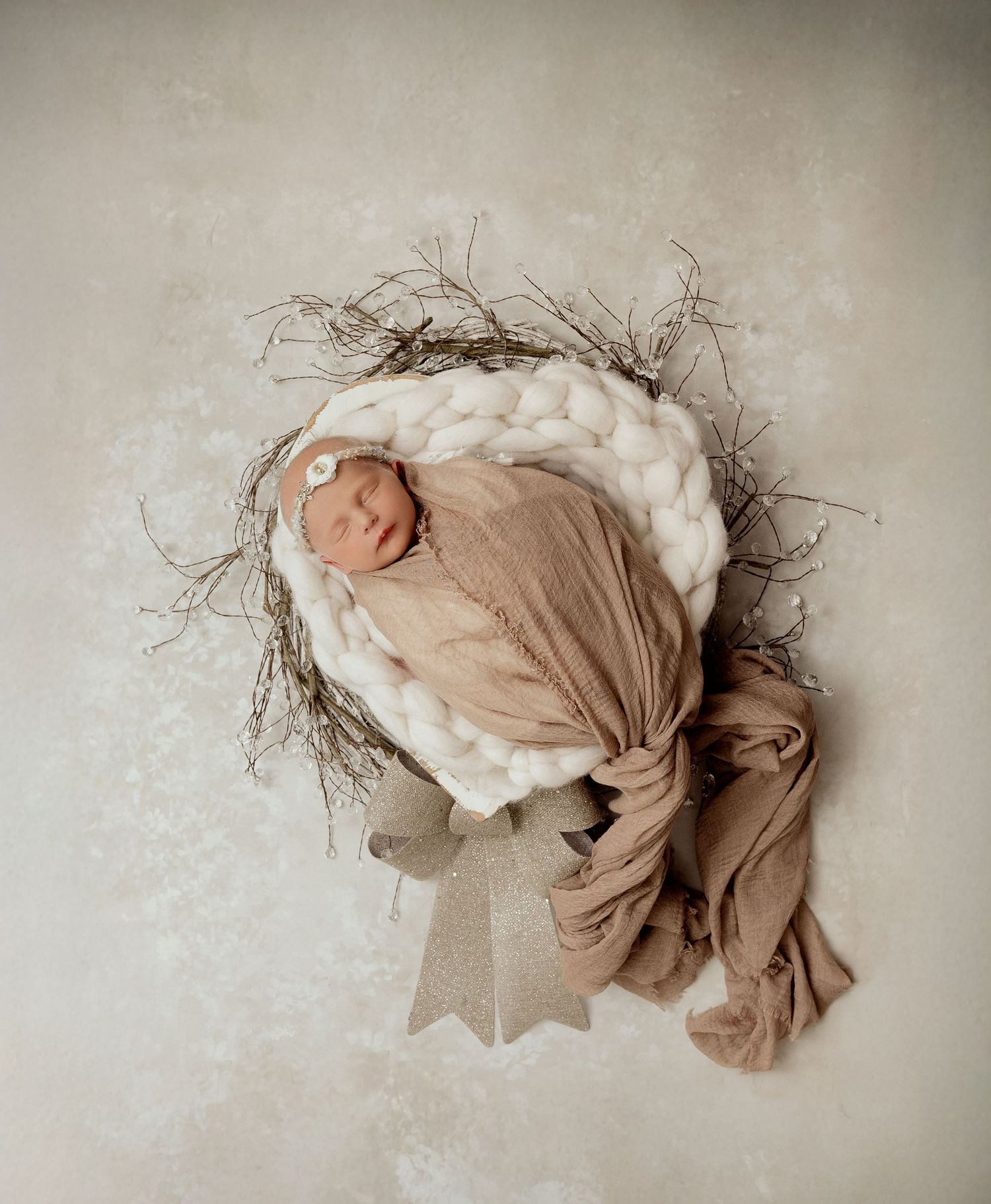 Newborn wrapped in beige fabric resting on chunky knit blanket with rustic twig decor.