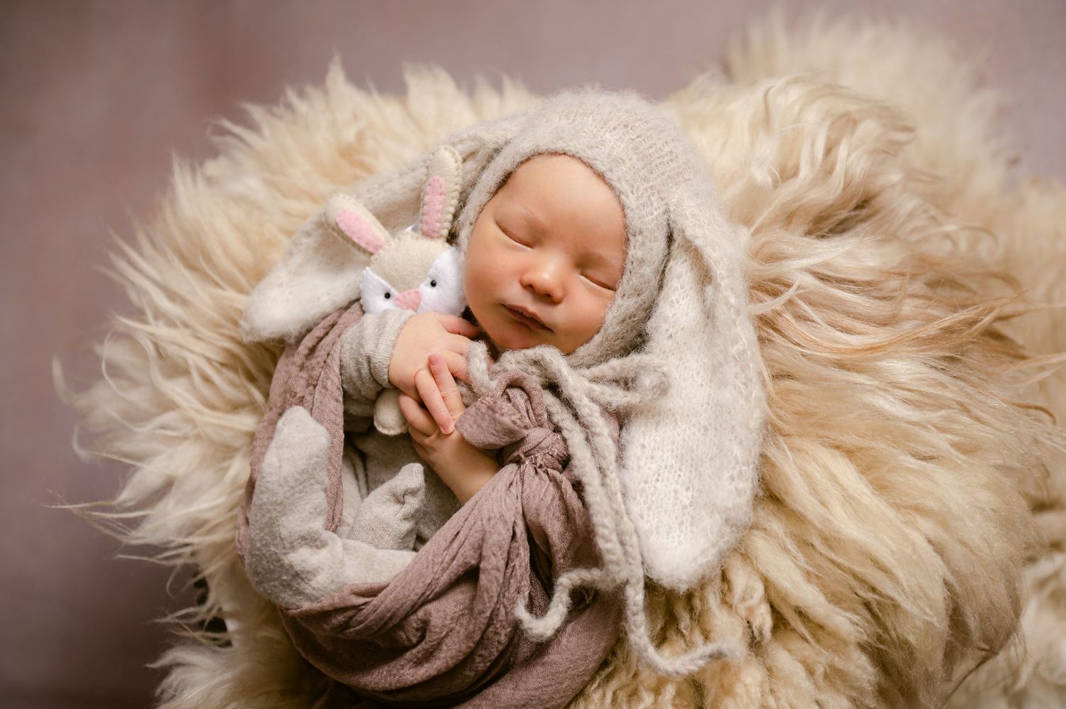 Sleeping baby wrapped in gray blanket snuggles with plush bunny on fluffy white fur backdrop.