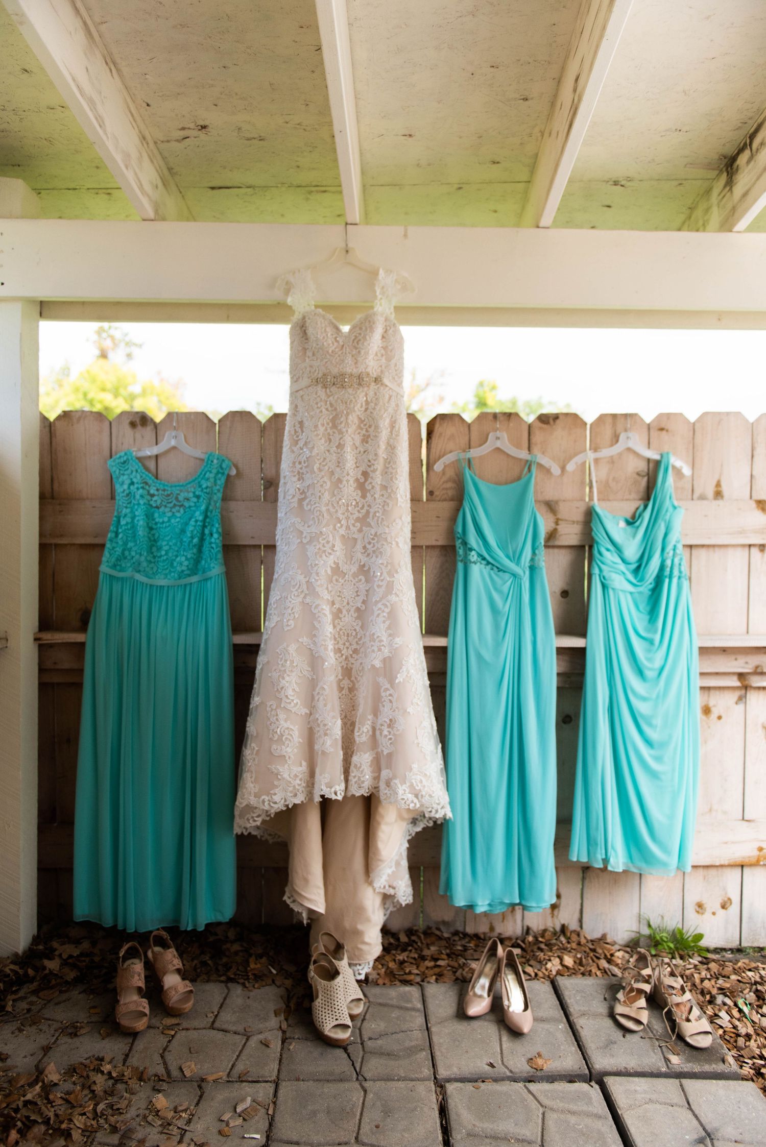 Wedding dresses and turquoise bridesmaid gowns hanging on a wooden fence with shoes below.