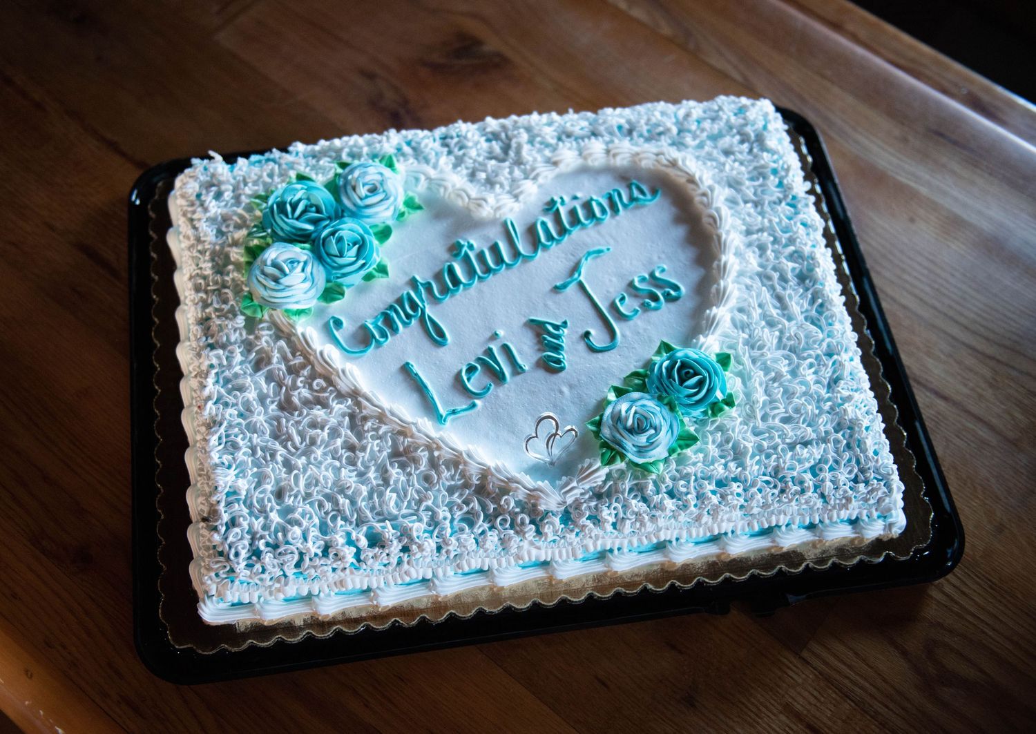 A white frosted wedding cake with teal flowers and congratulatory text displayed on a wooden table. Rum River Barn Milaca Minnesota Weddings