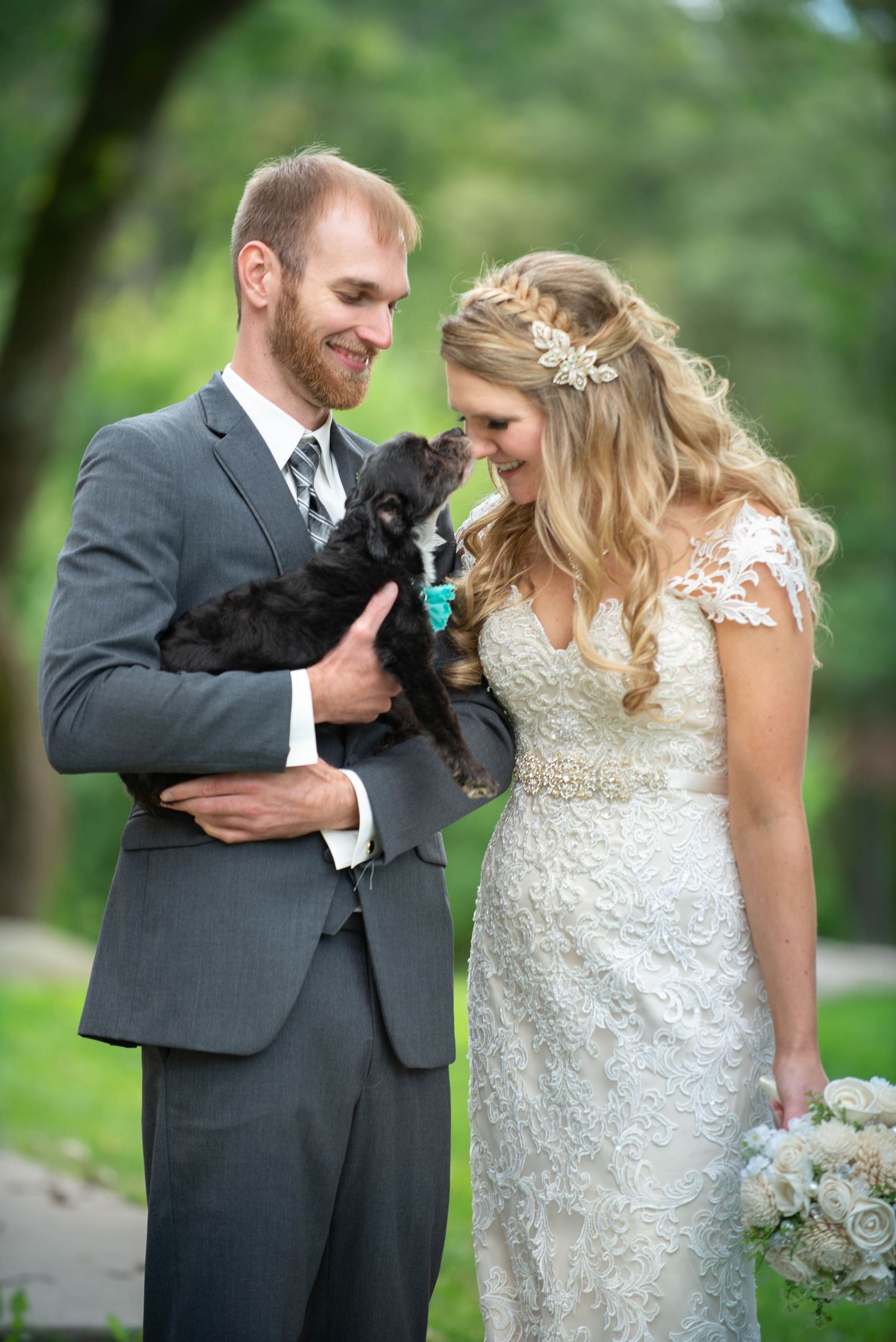 A couple in wedding attire shares a sweet moment with their small black dog in an outdoor garden setting. Rum River Barn Milaca Minnesota We