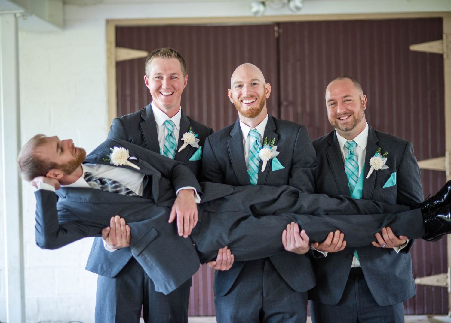 Groomsmen in matching tuxedos and turquoise ties playfully carry someone in a fun wedding party portrait.