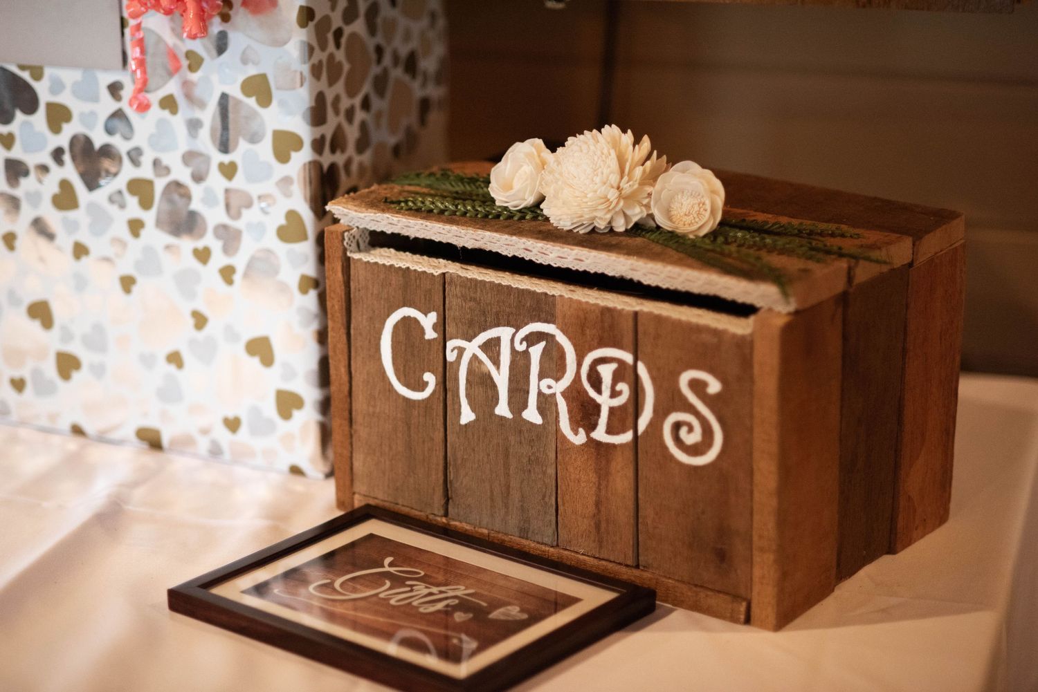 A rustic wooden card box decorated with white lettering and flowers sits on a table at a wedding reception.