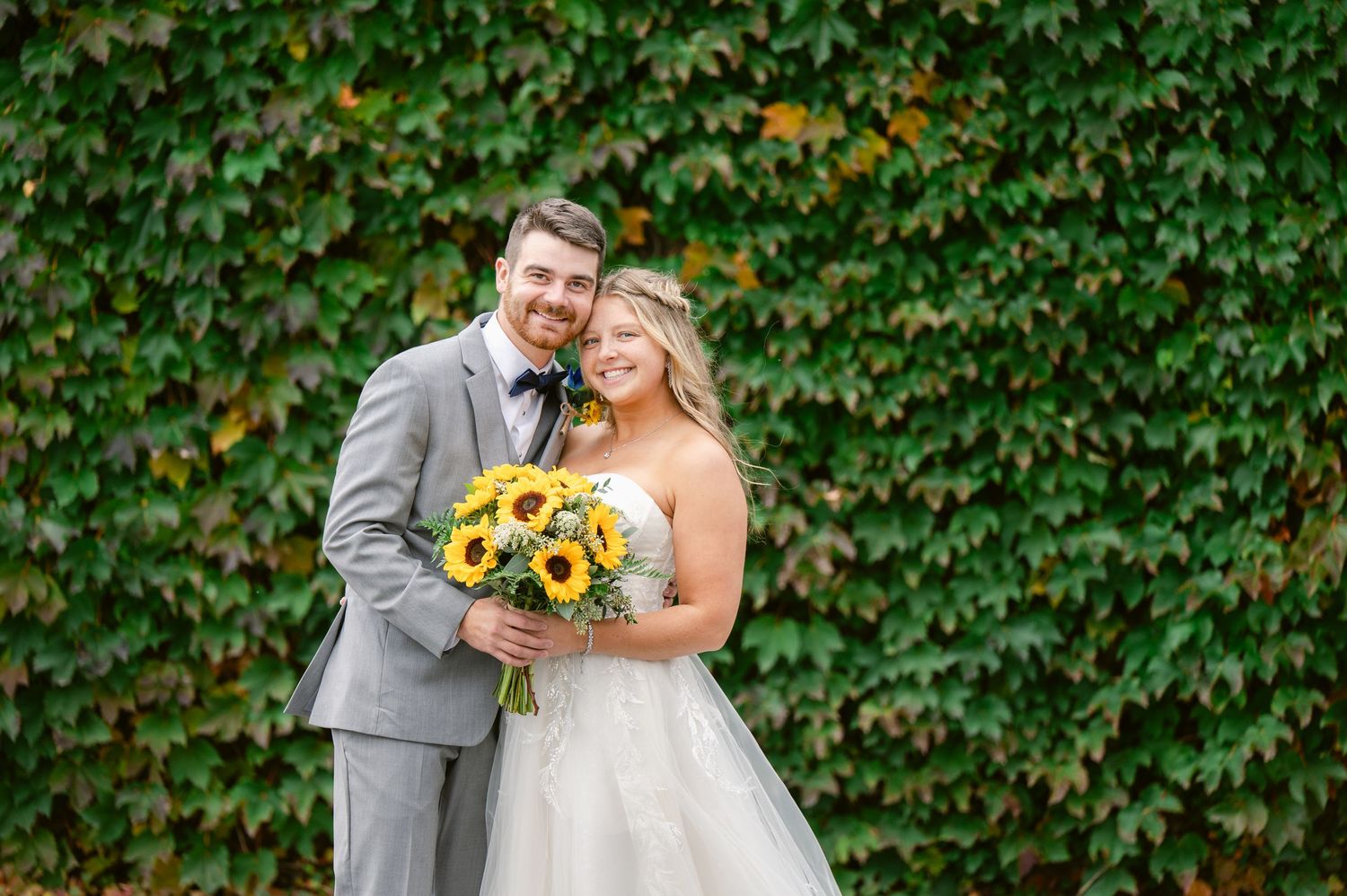 Couple poses with sunflower bouquet against ivy covered wall during wedding photoshoot.
