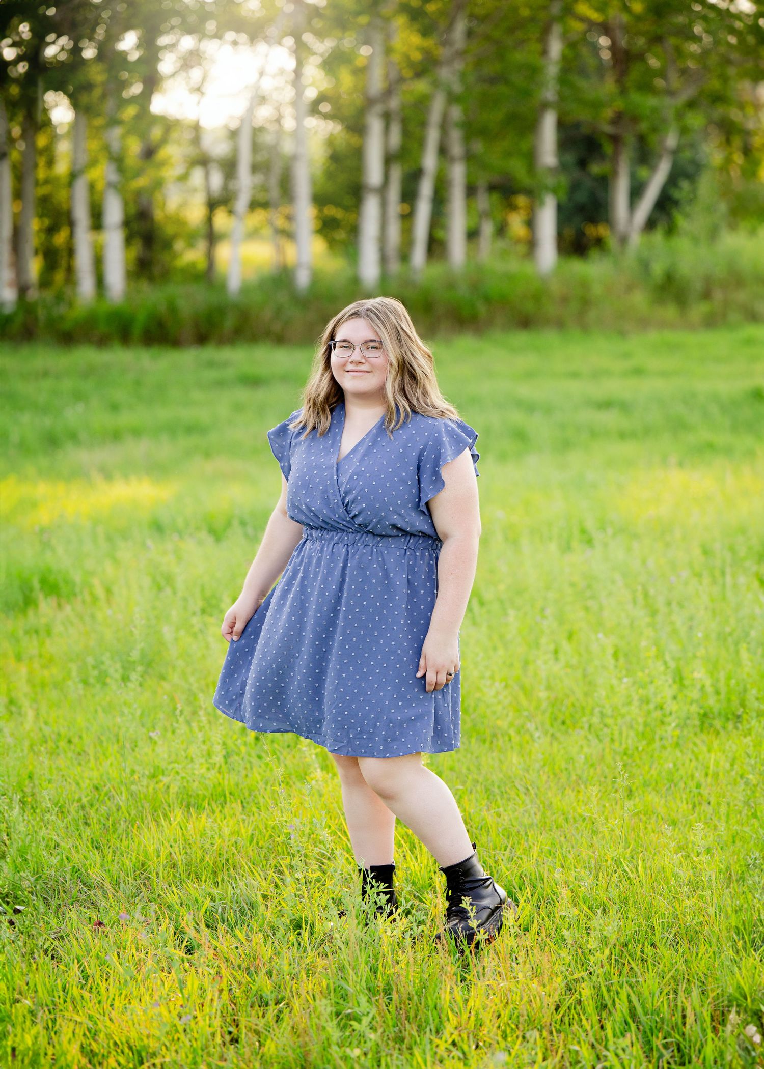 Senior Portrait in Foreston MN in a blue polka dot dress and black boots stands in a sunny meadow with birch trees in the background.