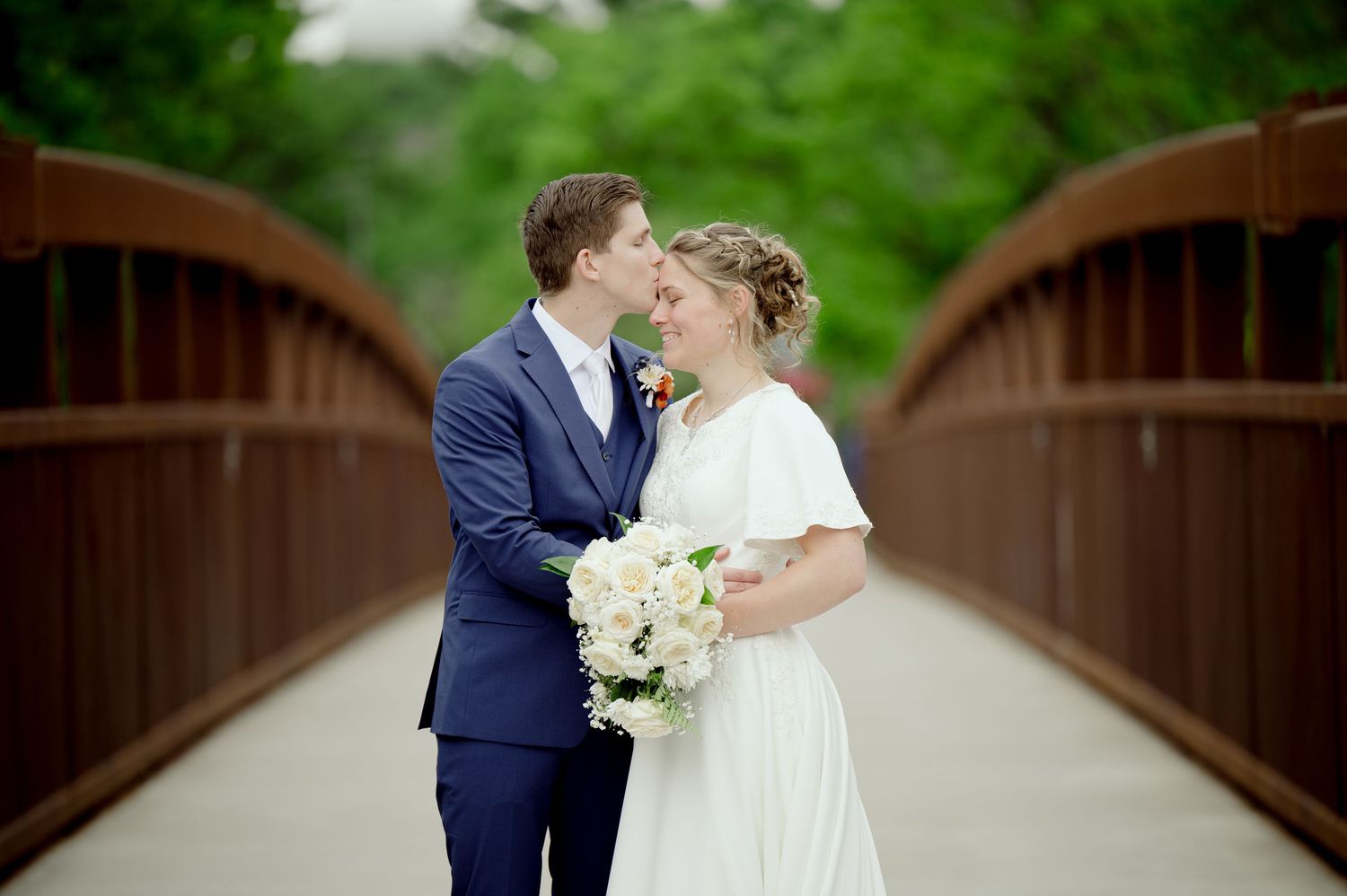 A couple kisses on a wooden bridge surrounded by lush greenery during their romantic outdoor wedding photoshoot.