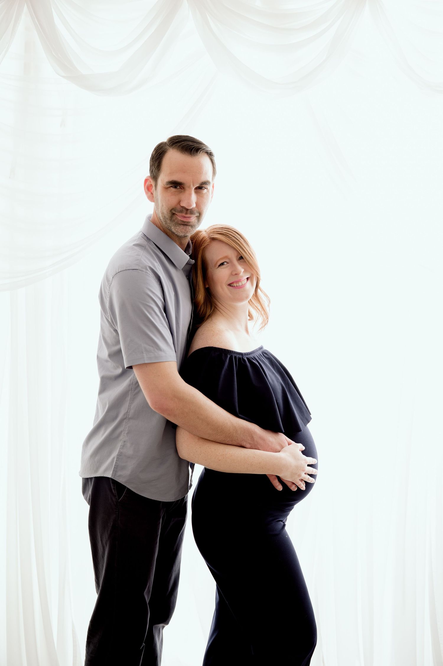 Expectant couple embrace in a tender maternity photoshoot with white curtains in the background.