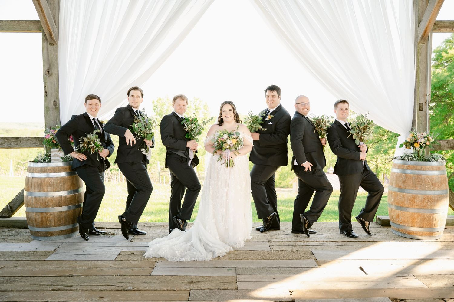 Wedding party poses in creative formation on wooden deck with white drapery backdrop.