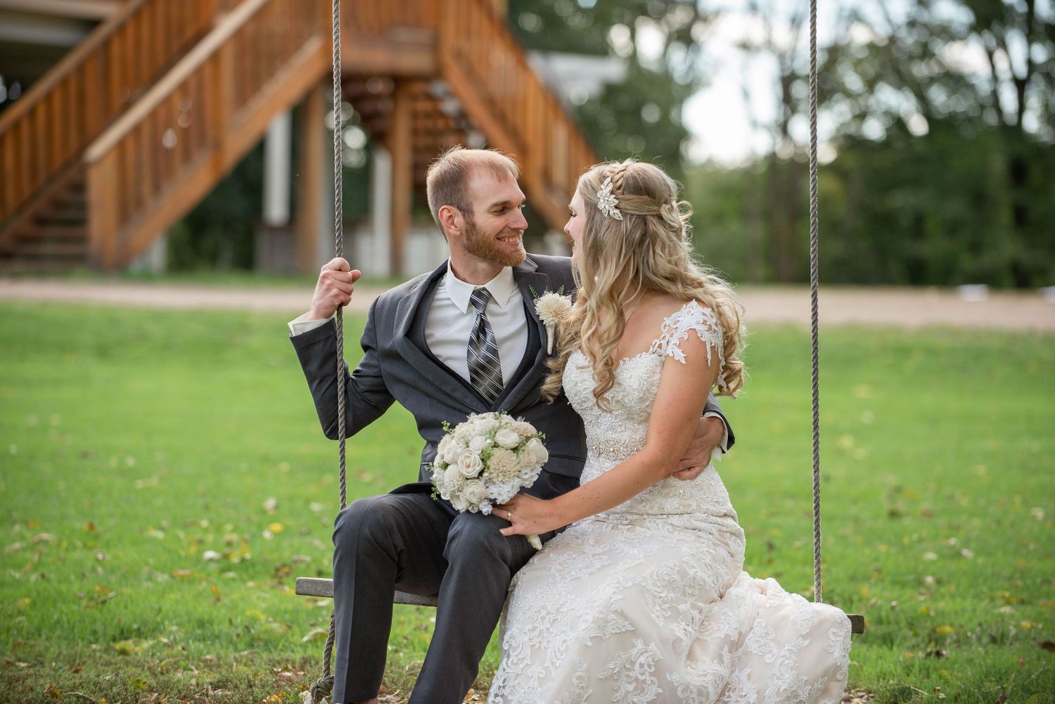 A newlywed couple sits together on a wooden swing in a garden setting with a bouquet of white flowers. Rum River Barn Milaca Minnesota Weddi