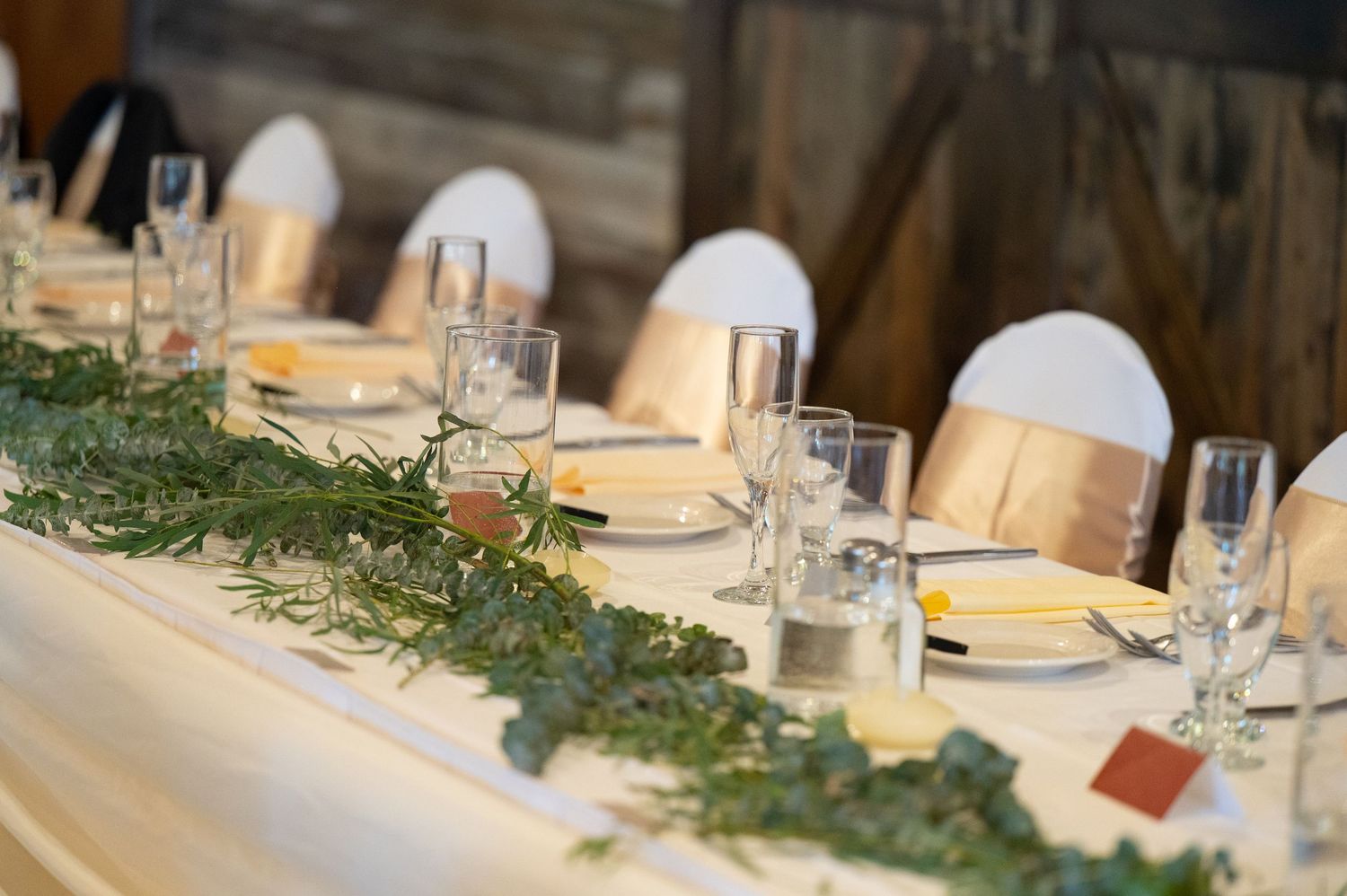 Rustic wedding reception table set with greenery garland, glasses and place settings in a barn venue.