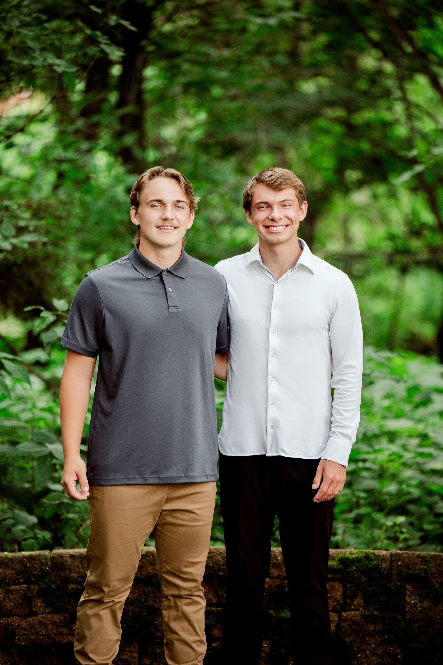 Two people stand together on a stone wall with a green forest backdrop wearing casual attire.