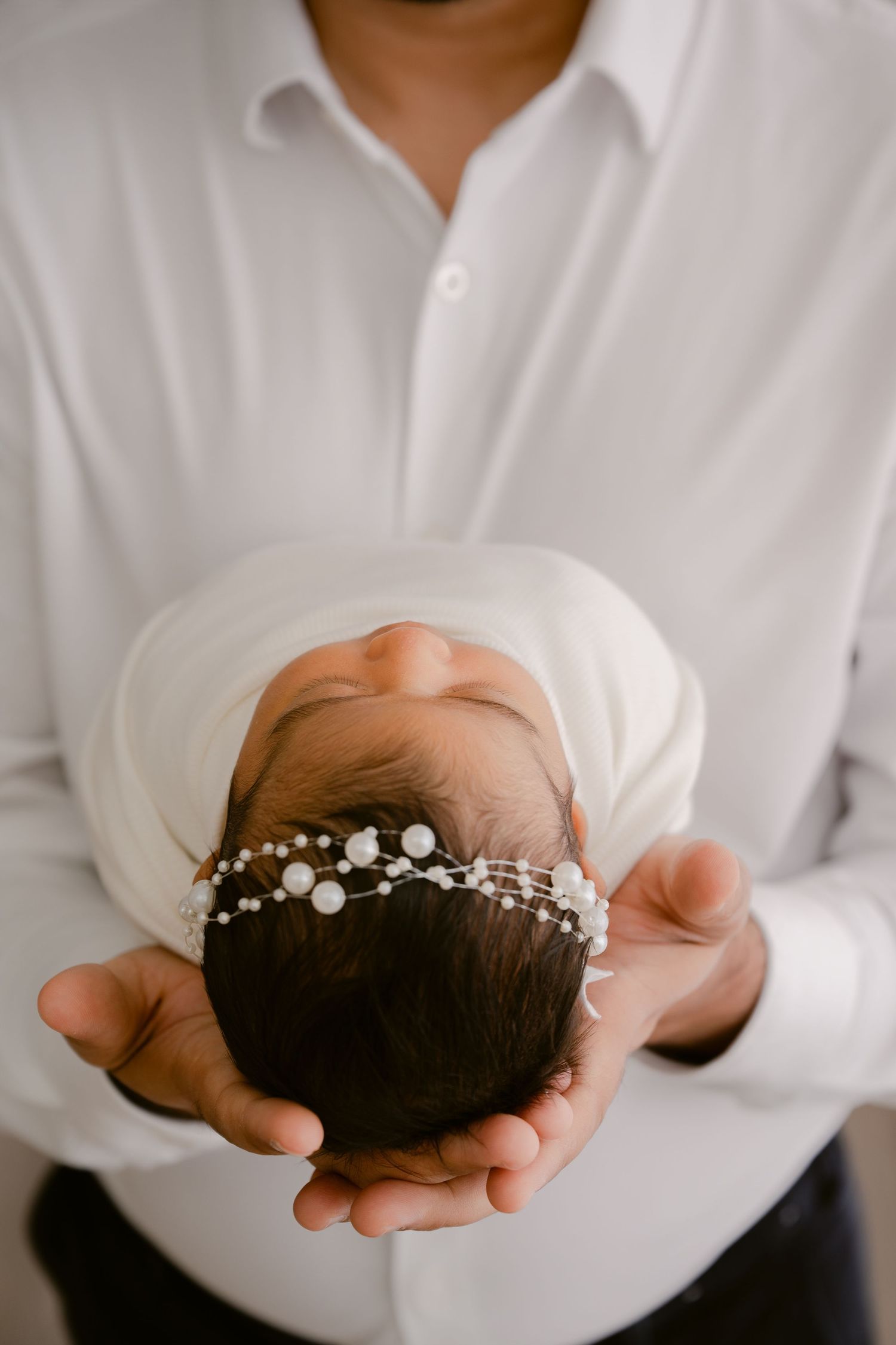Baby wearing white headband with pearls being cradled gently in adult hands.