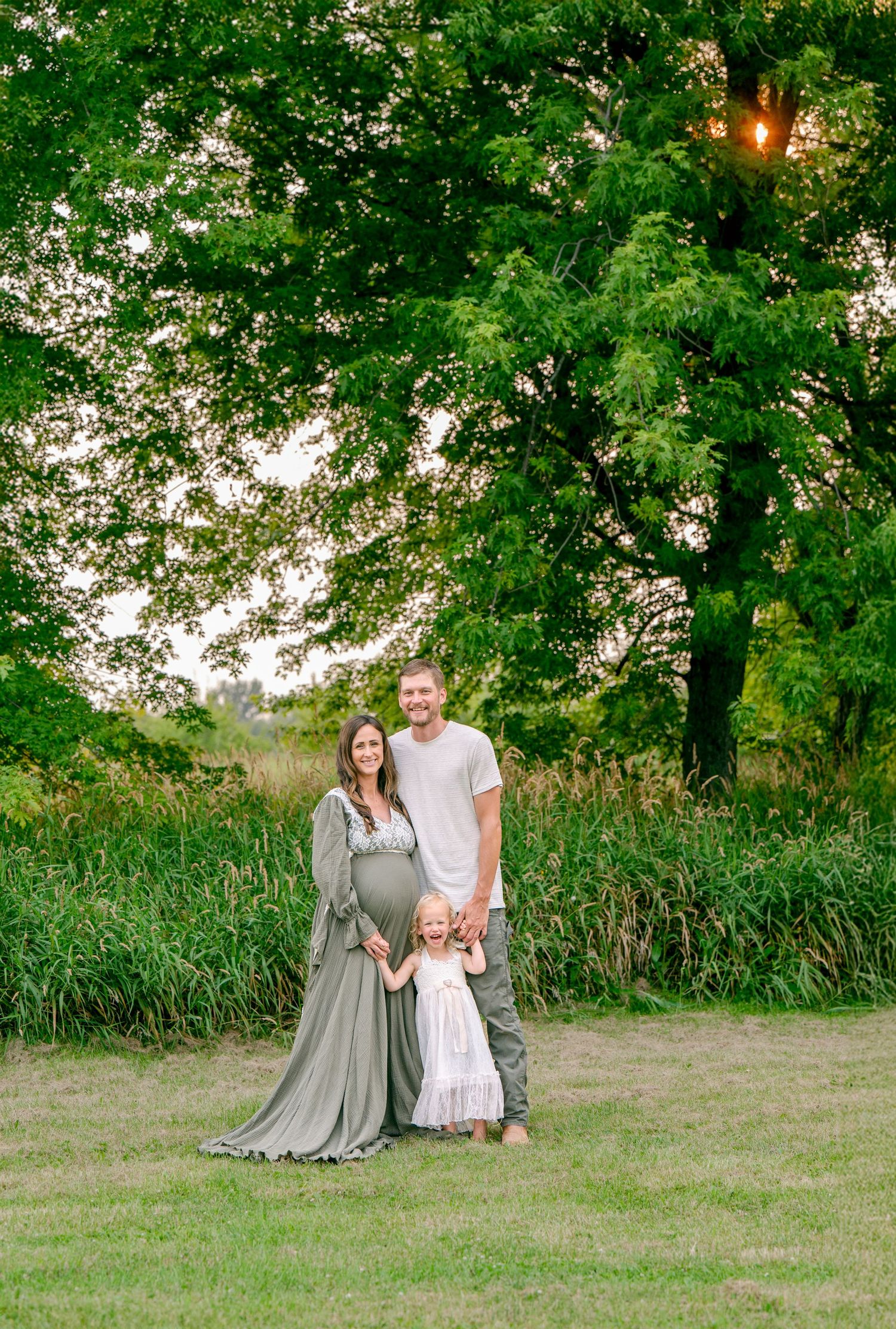 Family maternity photo showing parents and child in coordinated outfits standing beneath a large tree at sunset.