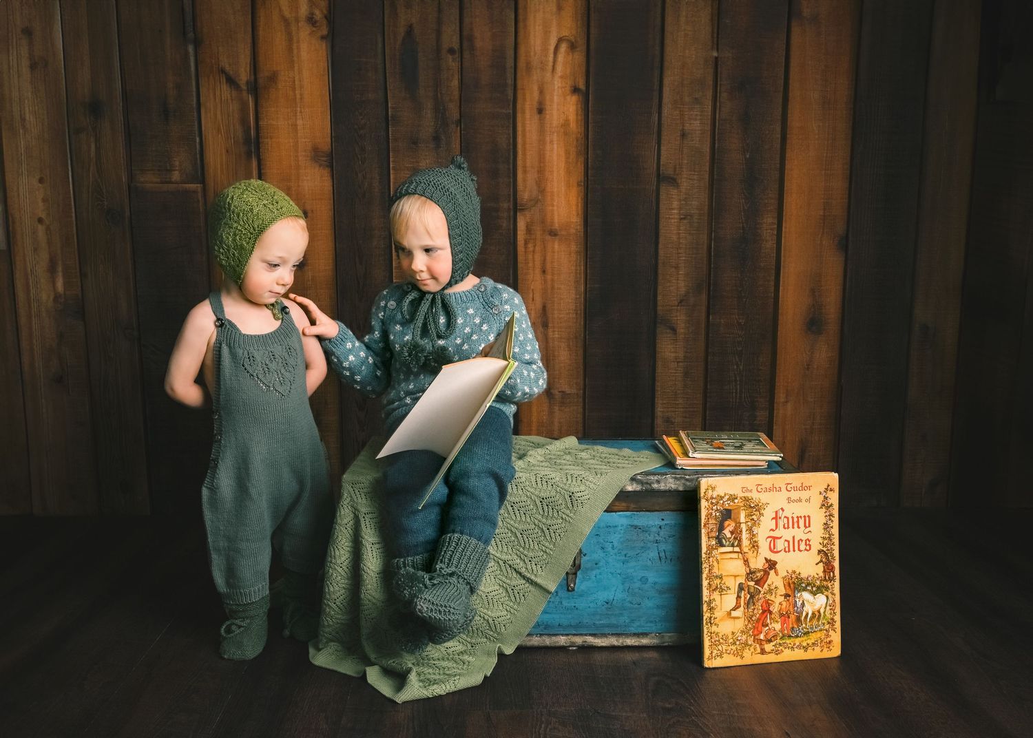 Two young ones looking at book while sitting on blanket against rustic wood wall.