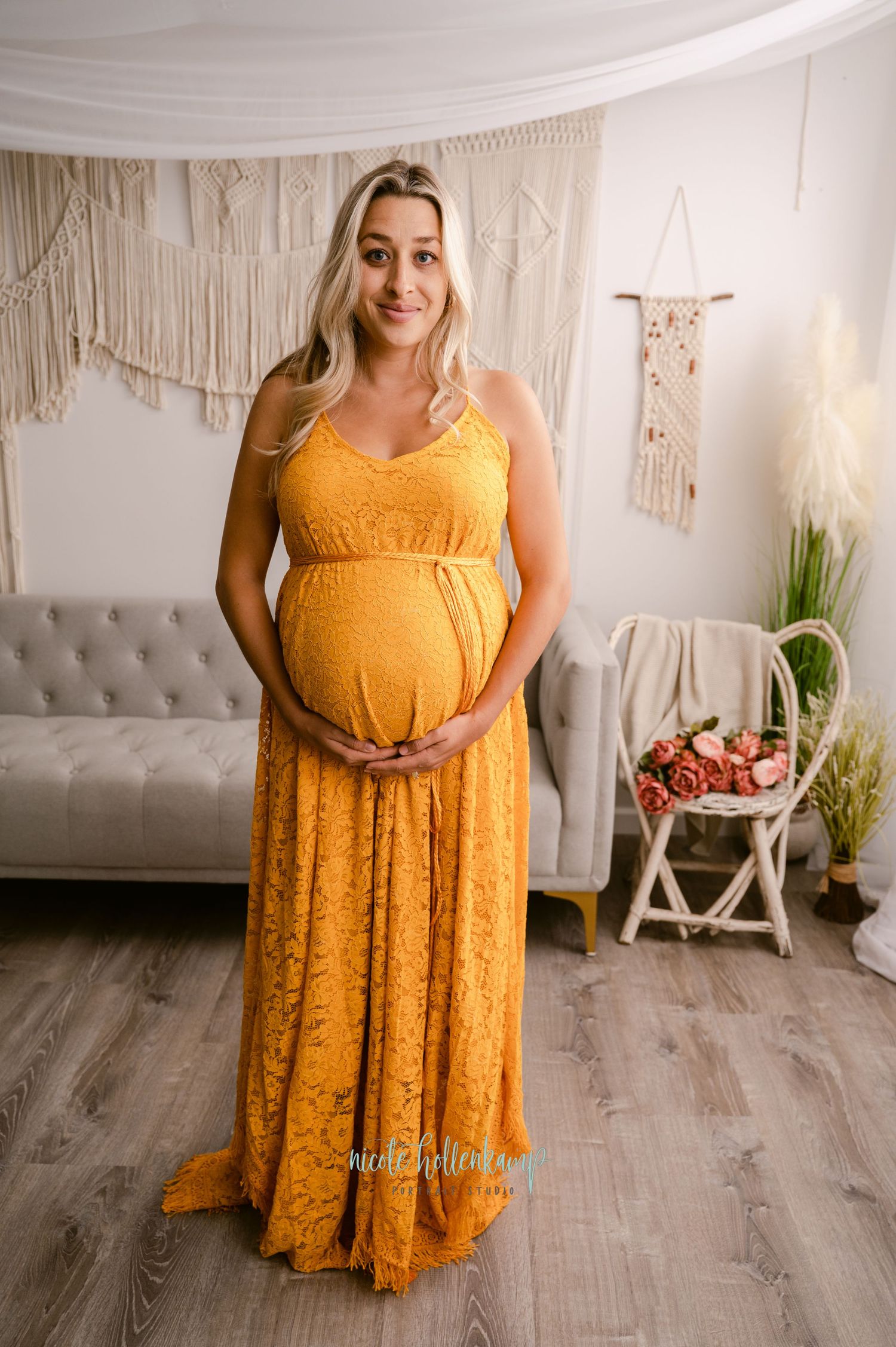 A pregnant person poses in a mustard yellow lace maxi dress against a boho-style backdrop with macrame wall hangings.