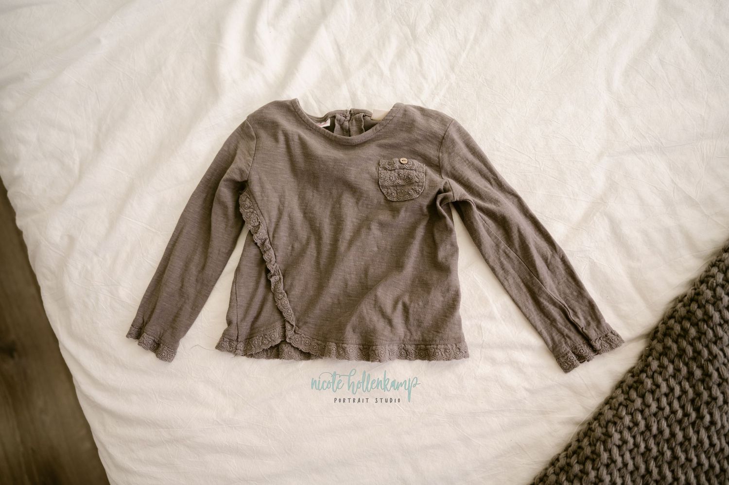 A vintage-style photograph of a taupe long-sleeved shirt laid flat on white bedding next to a textured woven mat.