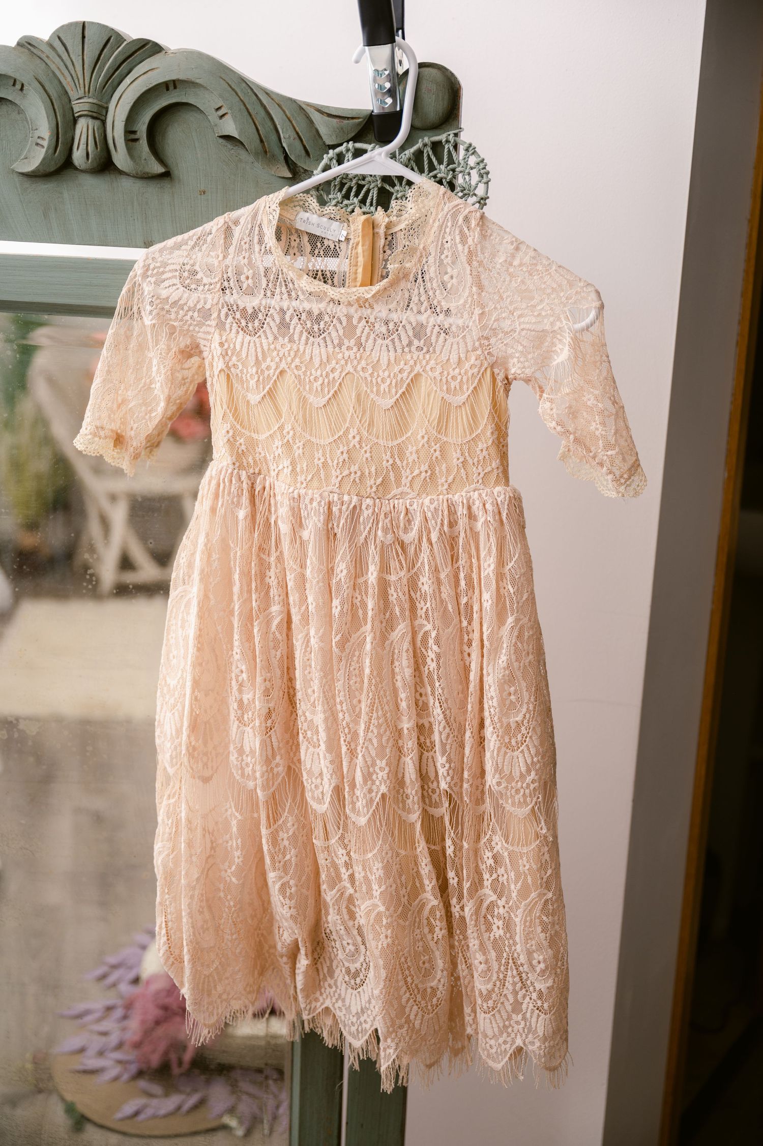 A delicate peach lace vintage-style dress with scalloped edges and short sleeves hangs on an ornate hanger against a wall.