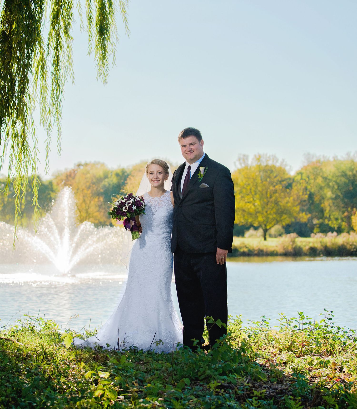 Newlyweds pose by a lake with fountain and weeping willow tree in background on sunny autumn day. Andover wedding at Magestic Oaks Golf Club