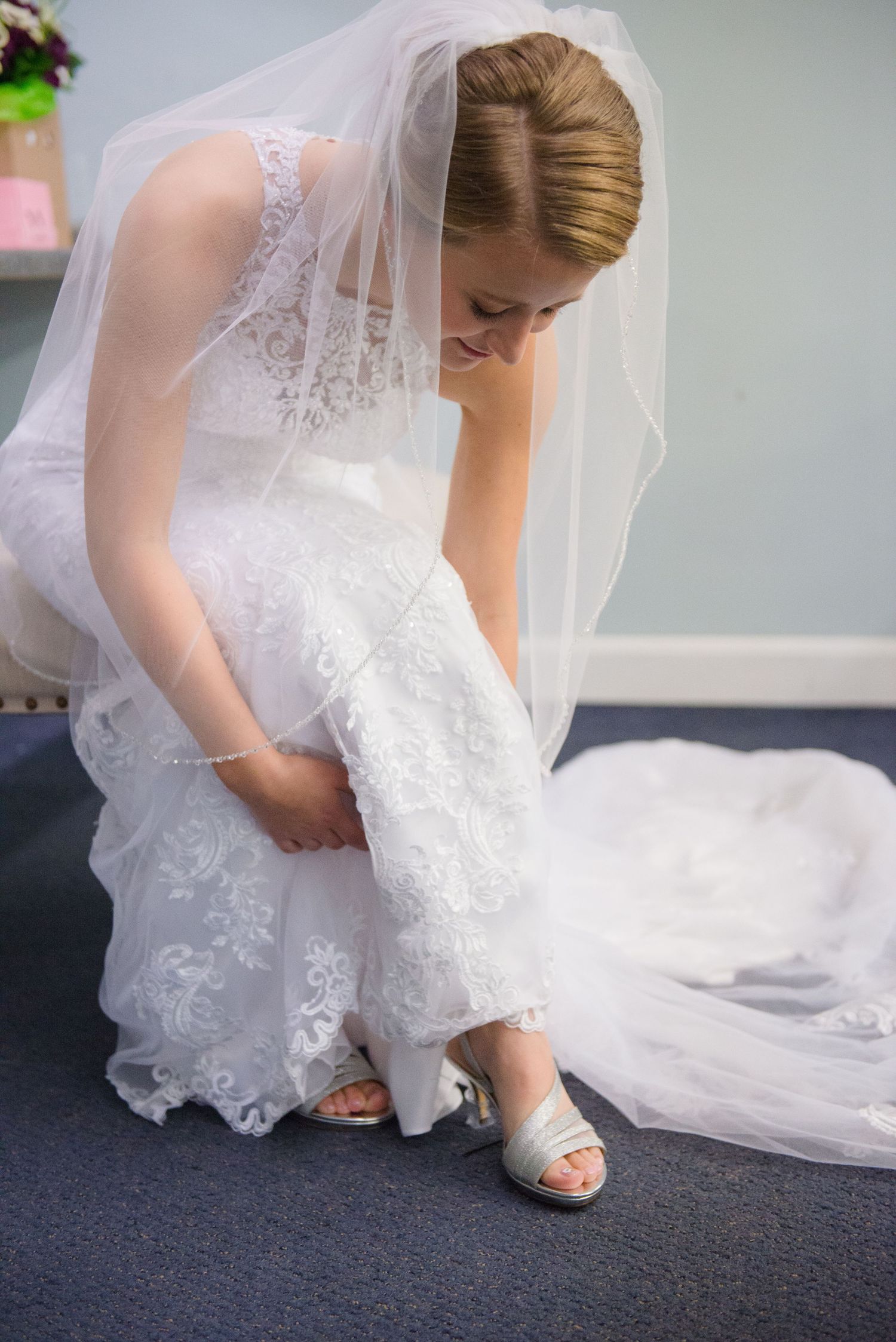 A close-up of someone adjusting their white wedding shoe while wearing a lace dress and veil. Andover wedding at Magestic Oaks Golf Club