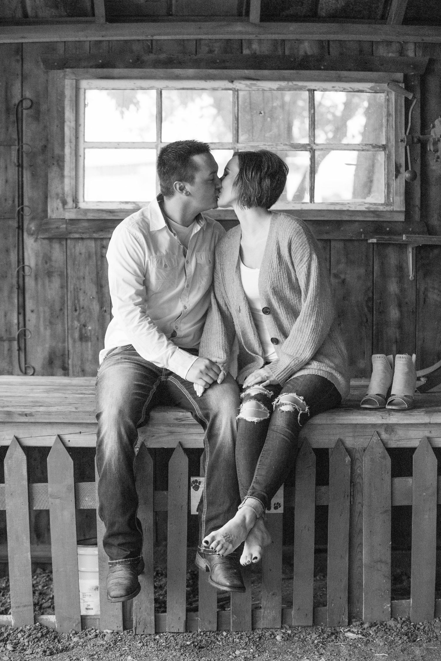 A romantic black and white photo of a couple sitting on a wooden fence by a rustic window, sharing an intimate moment.