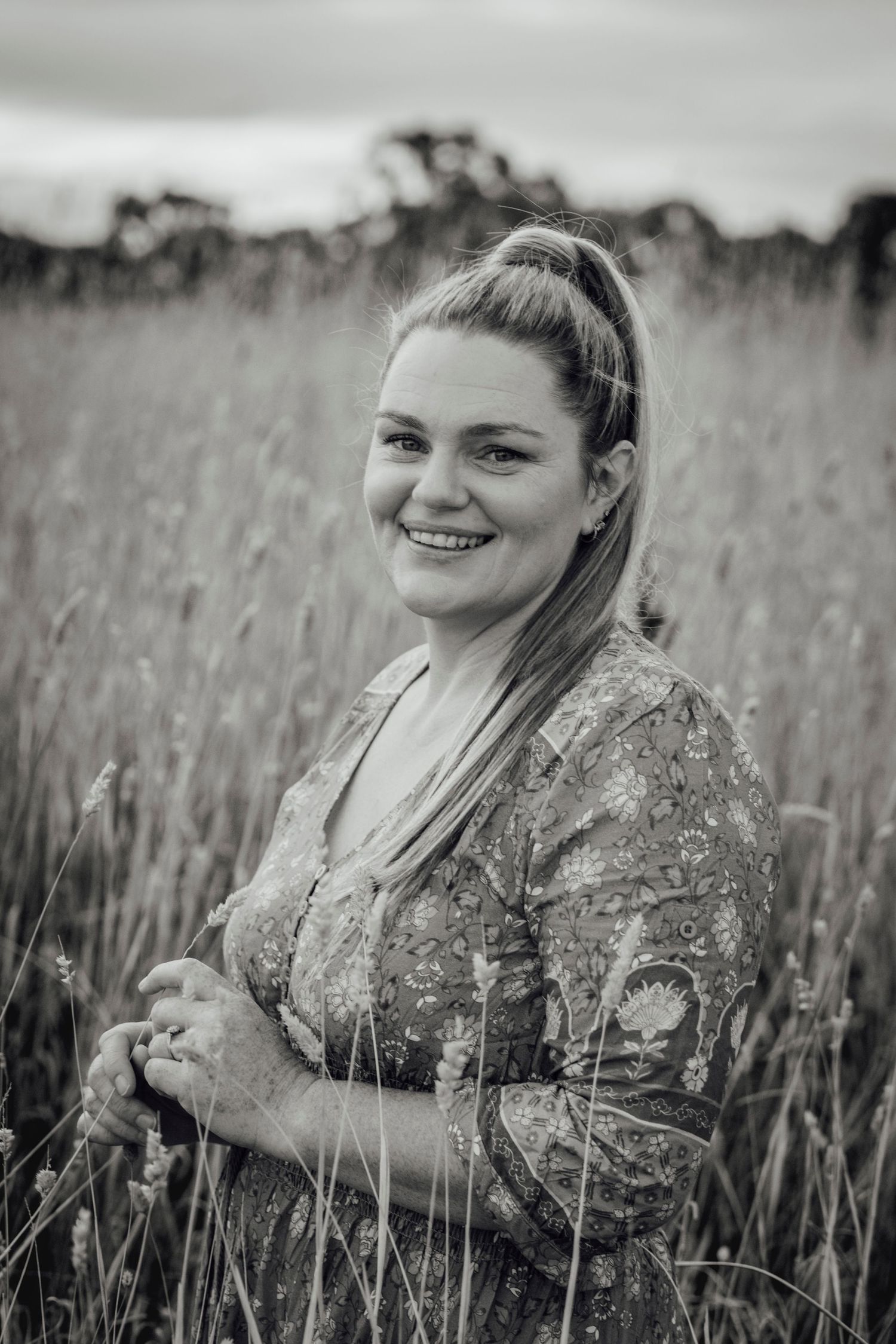 Black and white portrait of a smiling woman with a high ponytail wearing a floral dress standing in a tall grass field.