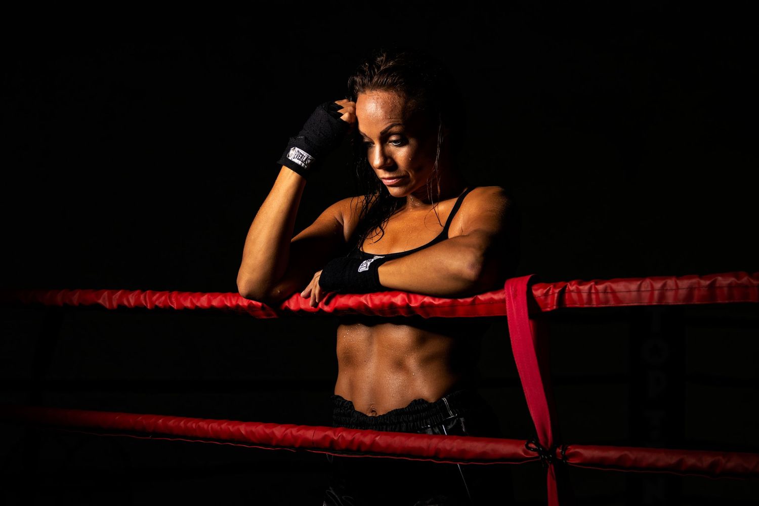 Dramatic low key photo of a boxer resting against red boxing ring ropes in dark lighting.