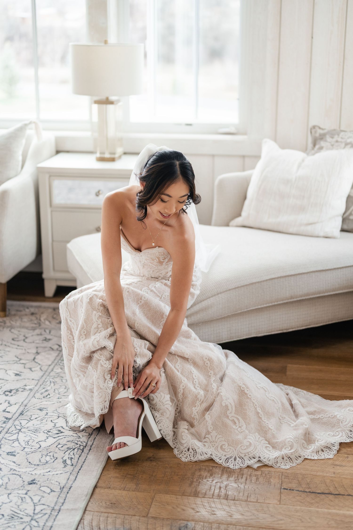 A bride in a delicate lace wedding dress adjusts her white heels while sitting in an elegant, light-filled room.
