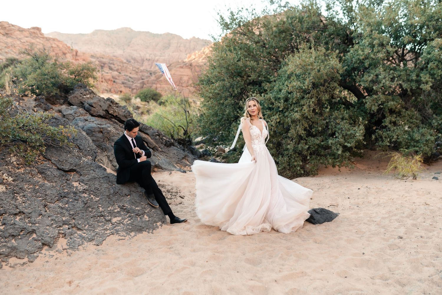 A couple sits together on desert rocks surrounded by desert landscape and greenery in a romantic outdoor setting.