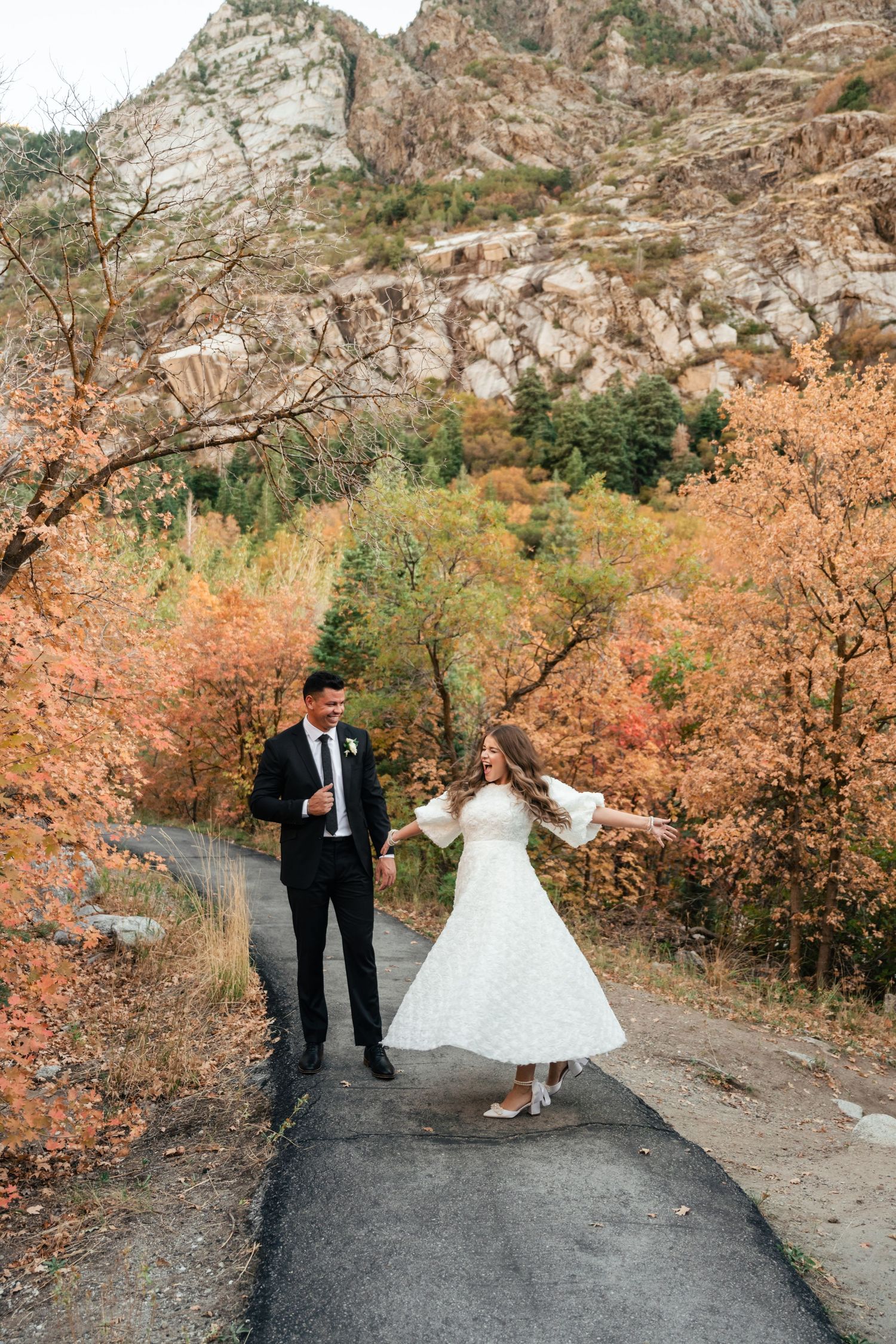 A couple in formal wedding attire dancing on a mountain path surrounded by autumn foliage and rocky cliffs.