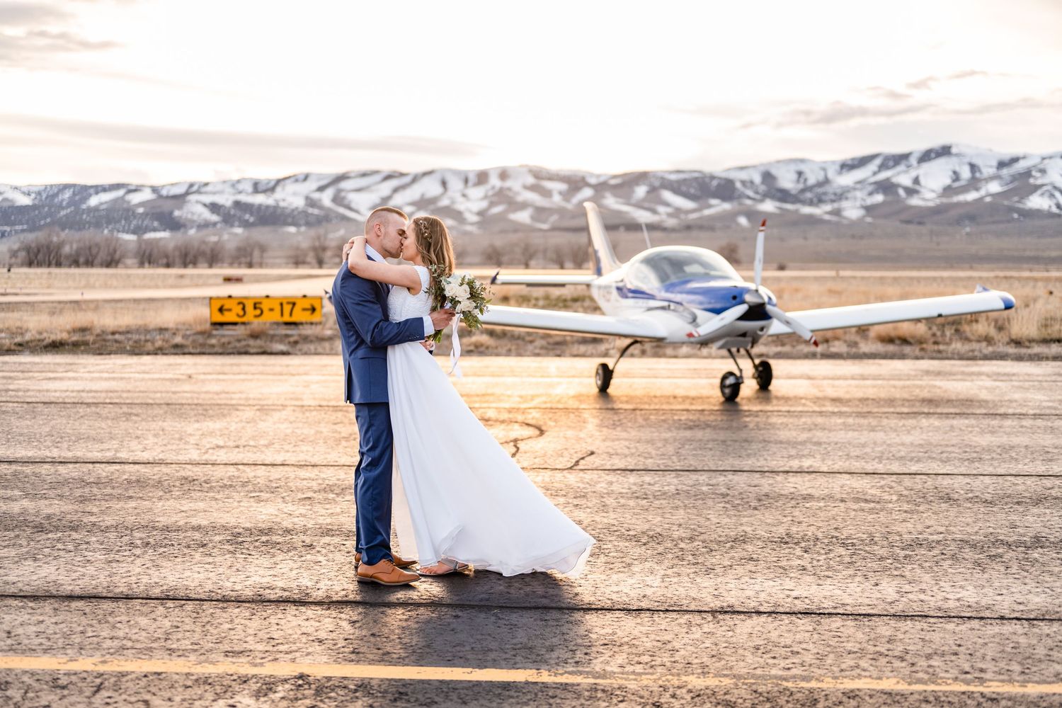 A couple embrace on an airfield tarmac with a small private plane and snow-capped mountains in the background.