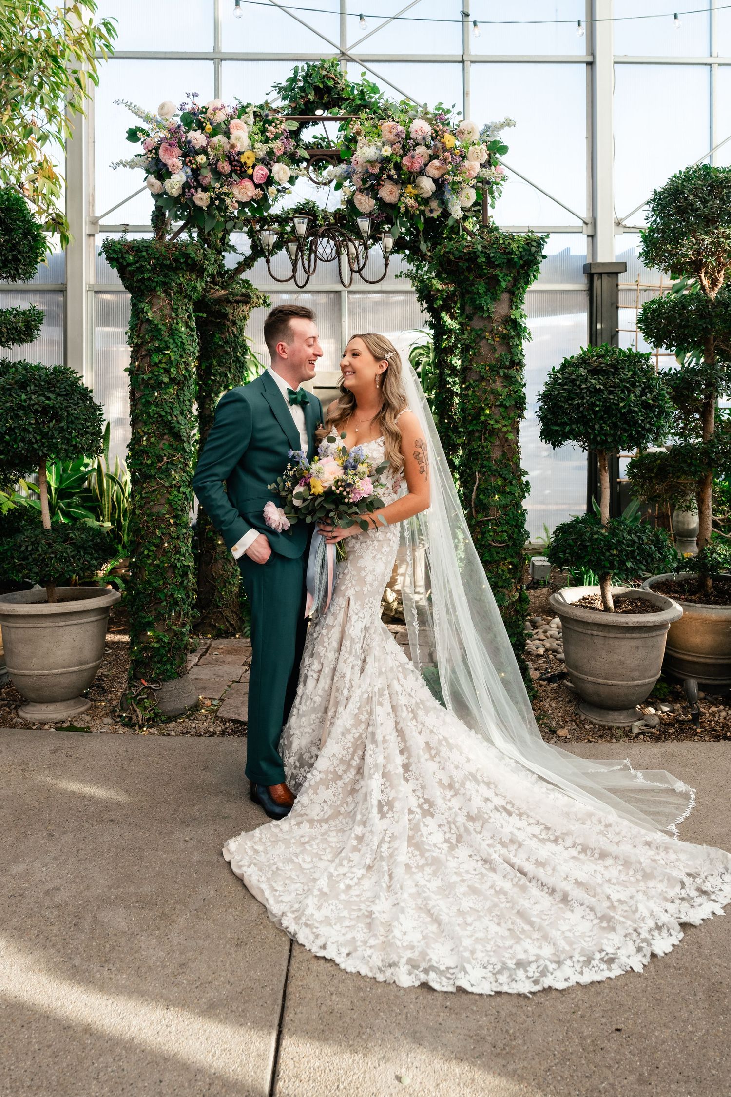 Wedding couple stands under floral arch in greenhouse with potted topiaries and trailing lace veil.