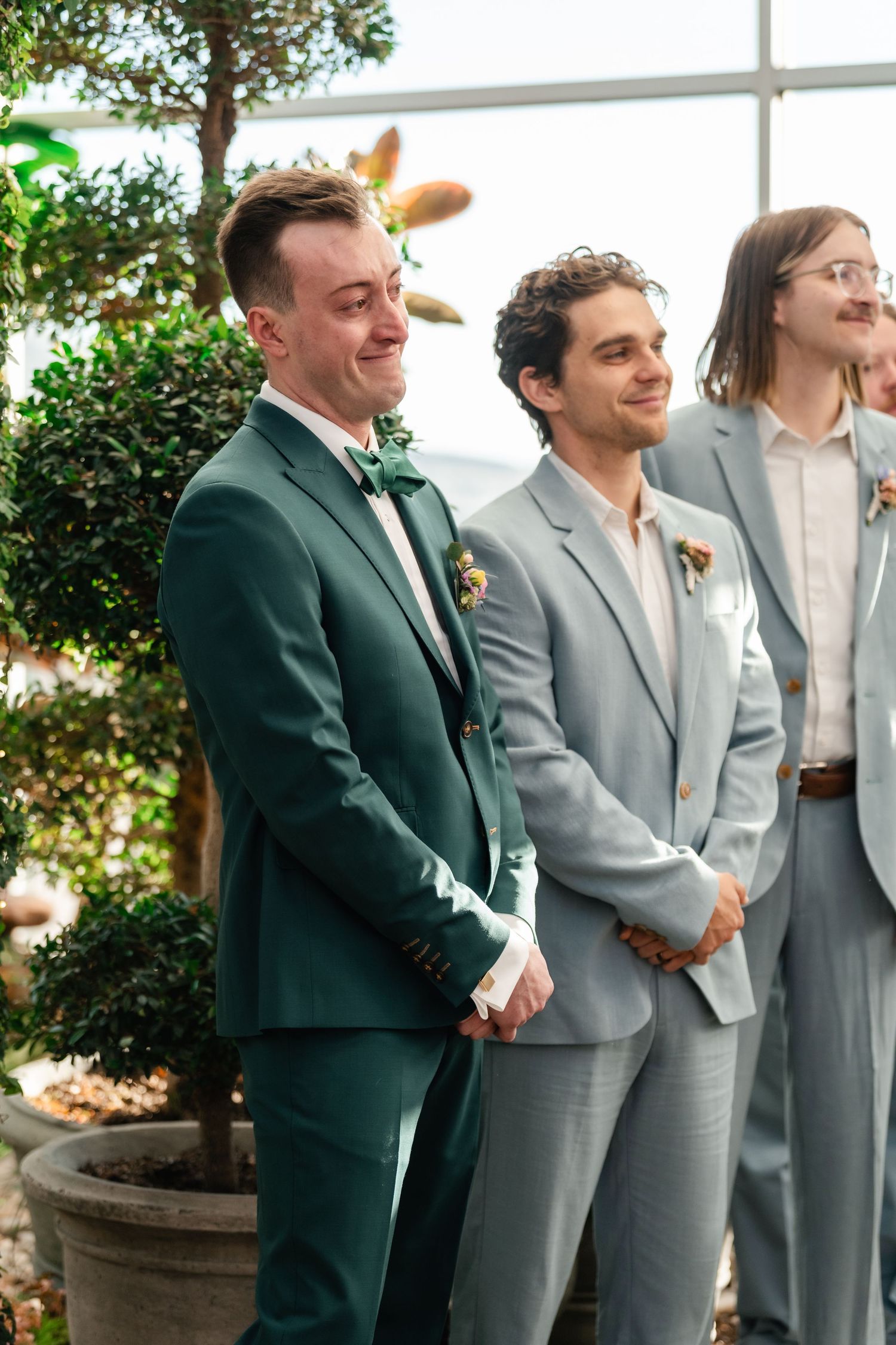 Three groomsmen stand together at a wedding ceremony wearing coordinated green and gray suits with boutonnieres.
