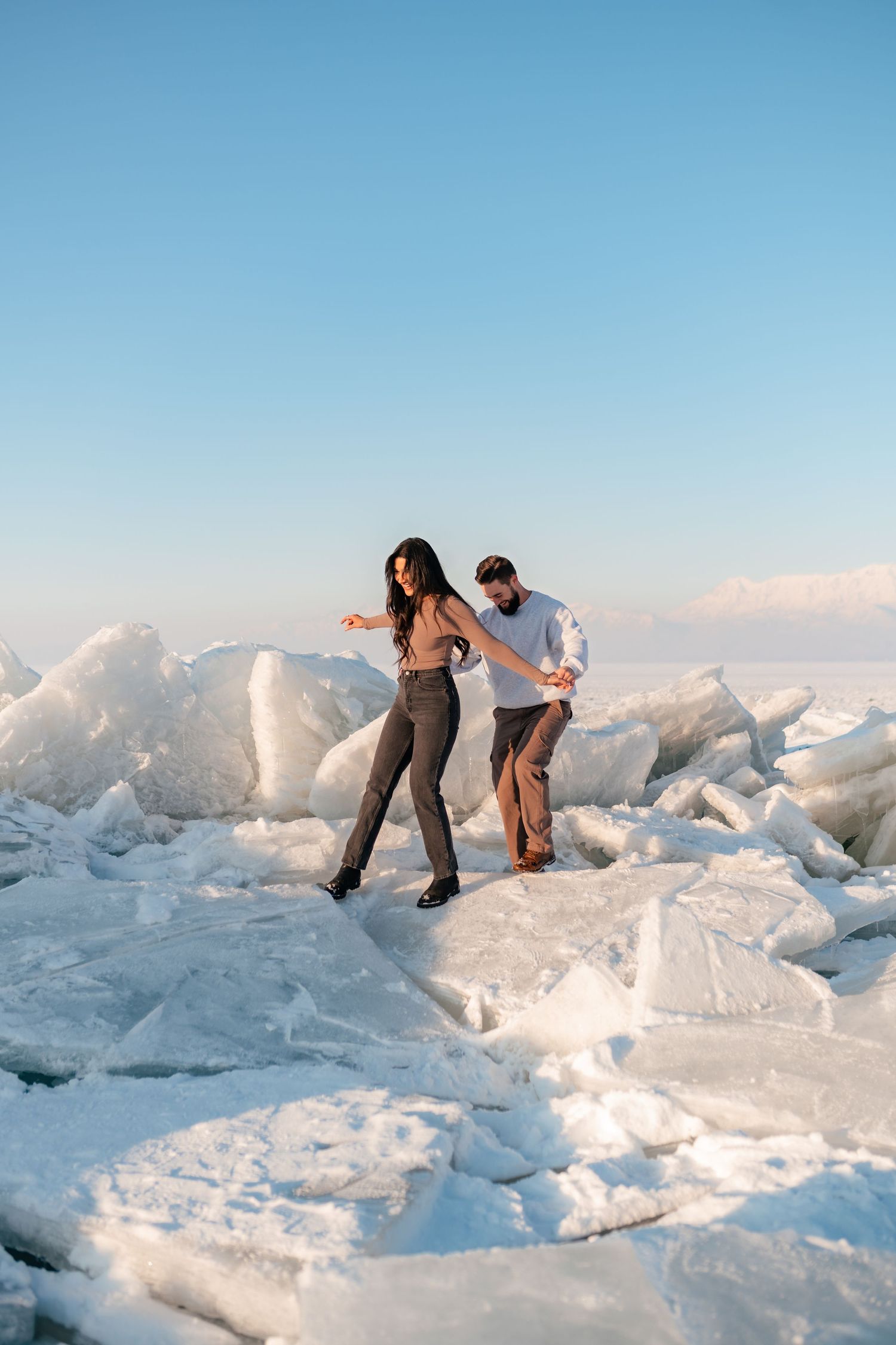 A couple walks carefully across jagged ice formations against a bright blue sky during sunset.