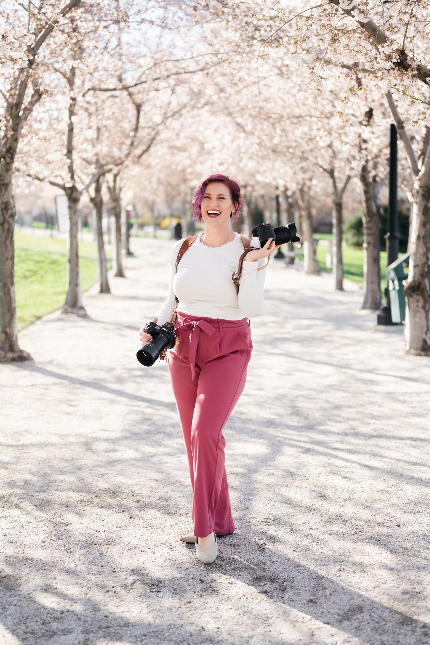 Photographer holding cameras walks along cherry blossom-lined path in spring wearing white top and pink pants.