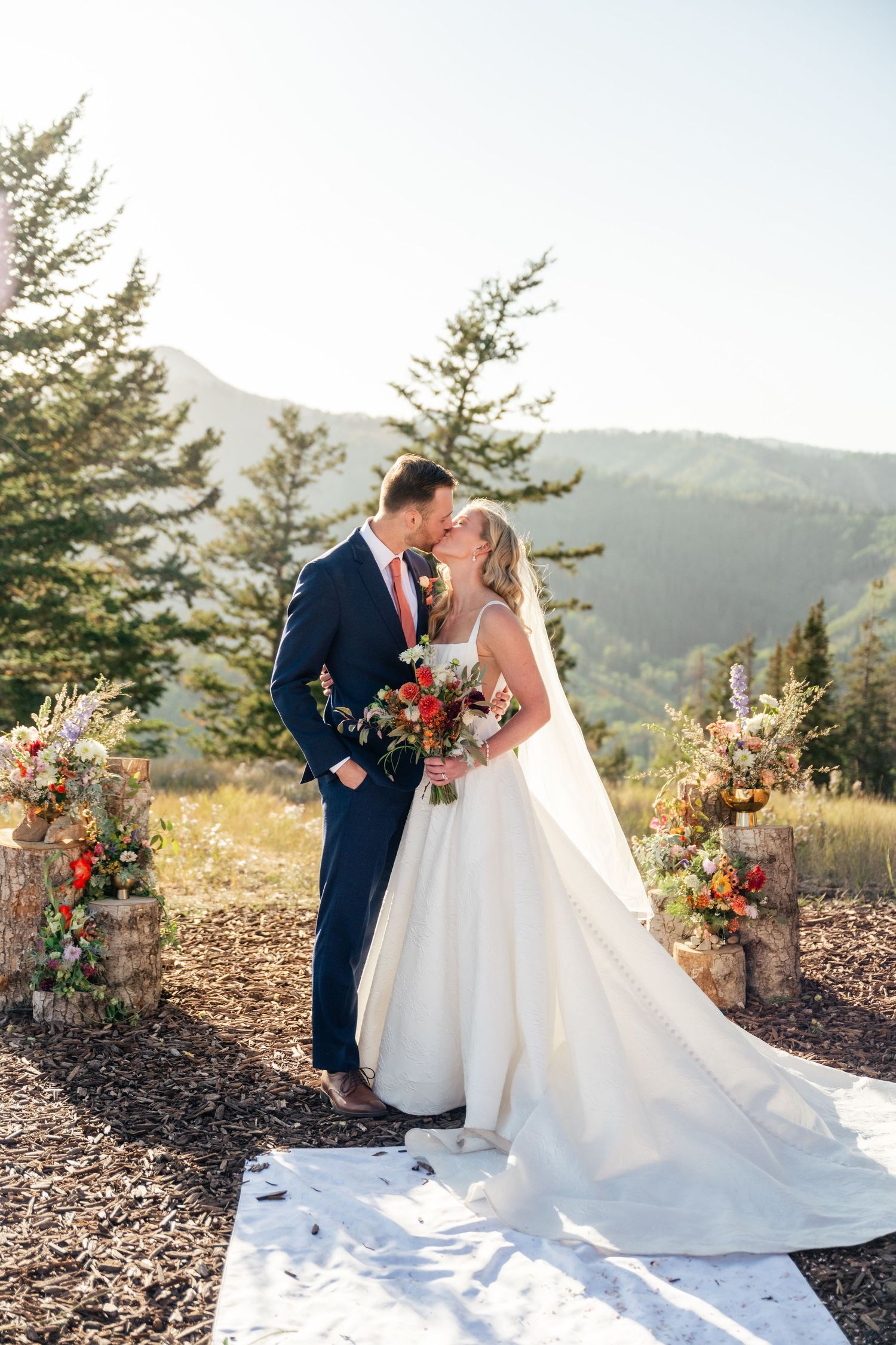 A couple in wedding attire share a romantic moment against a mountain backdrop with evergreen trees and floral arrangements.