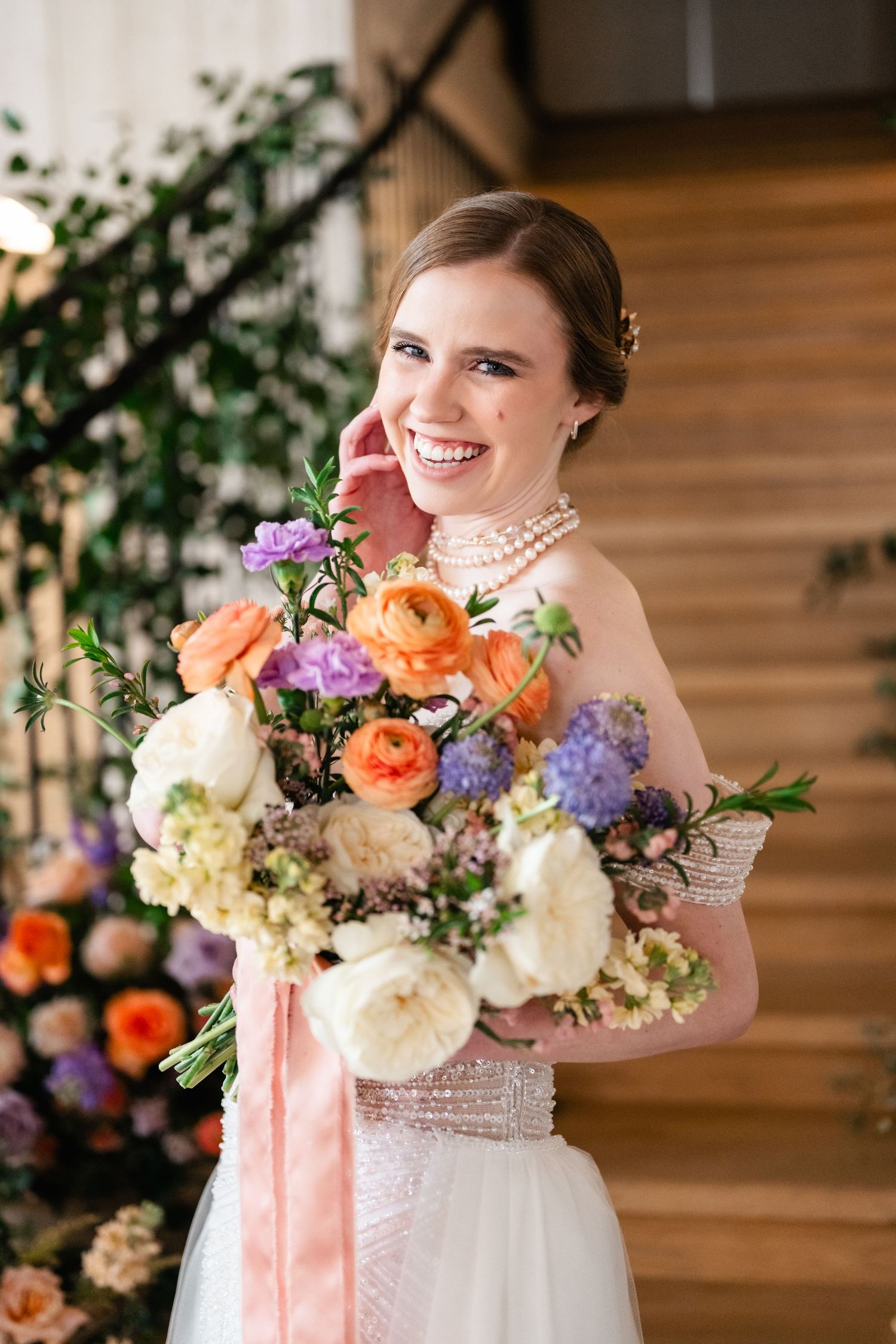 A bride in a white dress smiles while holding a vibrant bouquet of orange, purple and white flowers with silk ribbons.