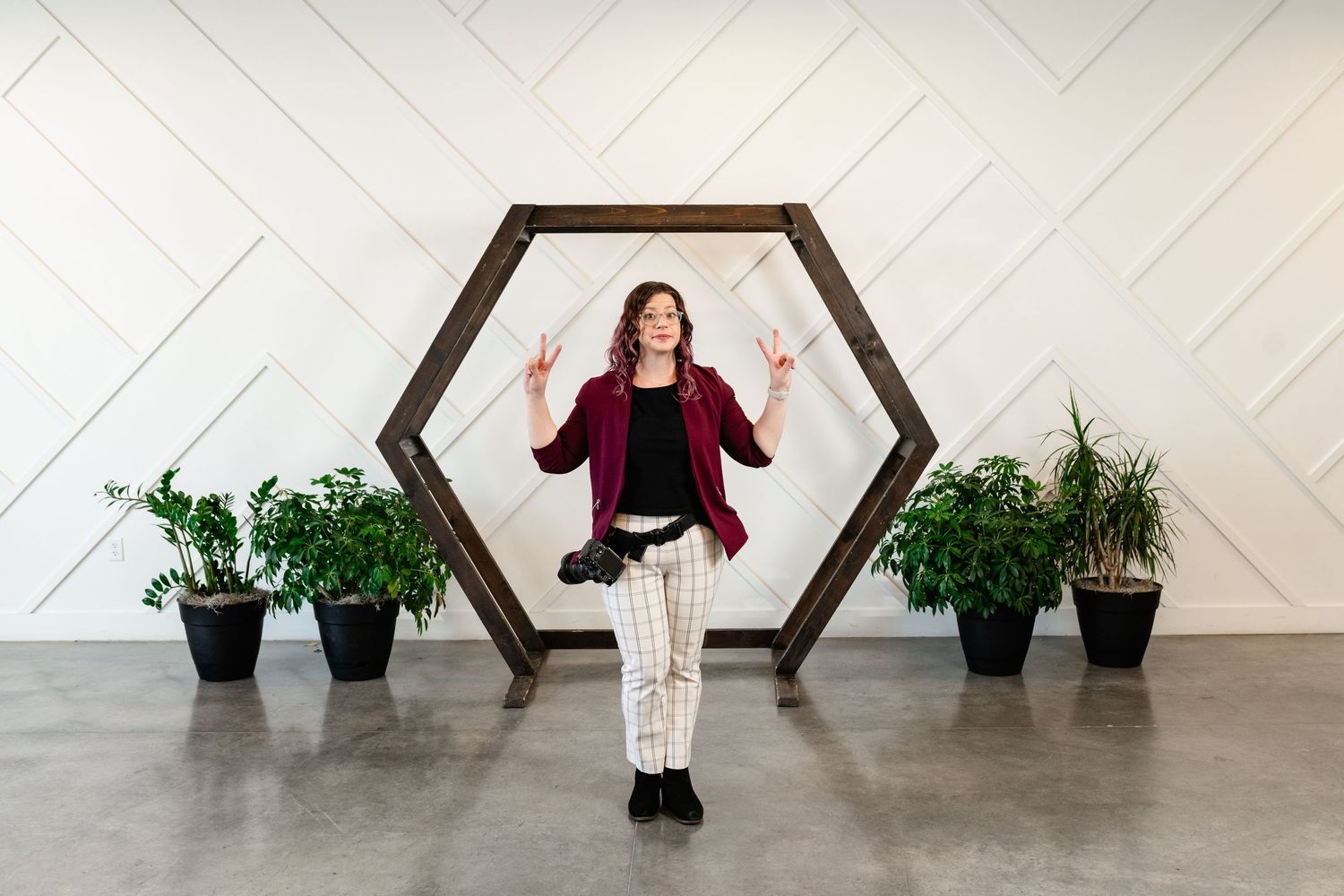 Someone poses with raised hands against a hexagonal wood frame with plants on either side against a white geometric wall.